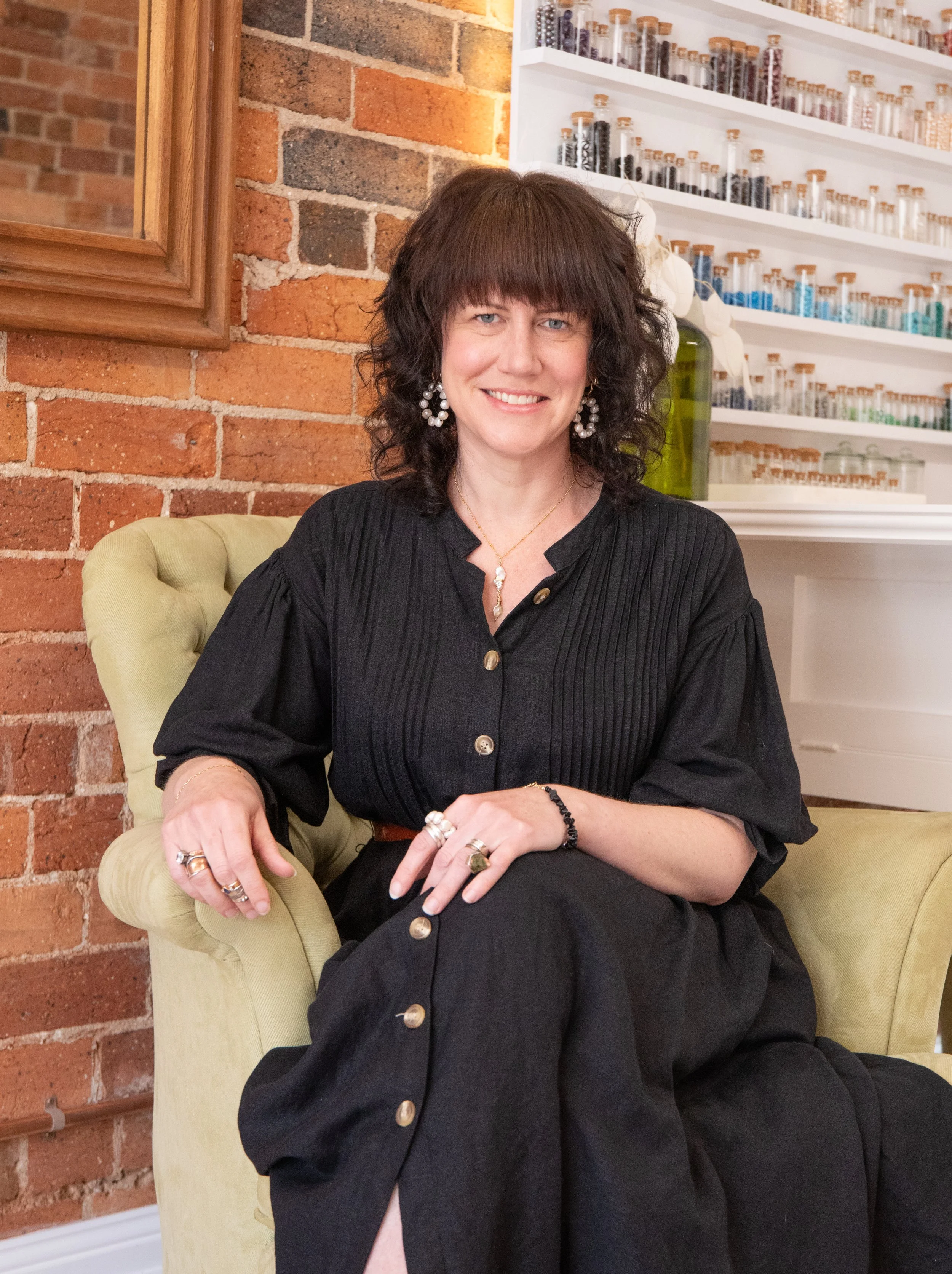 A woman with shoulder-length curly dark hair, wearing a black dress with gold buttons, sitting in a green armchair in front of a brick wall with a mirror and a white shelf filled with small glass bottles.