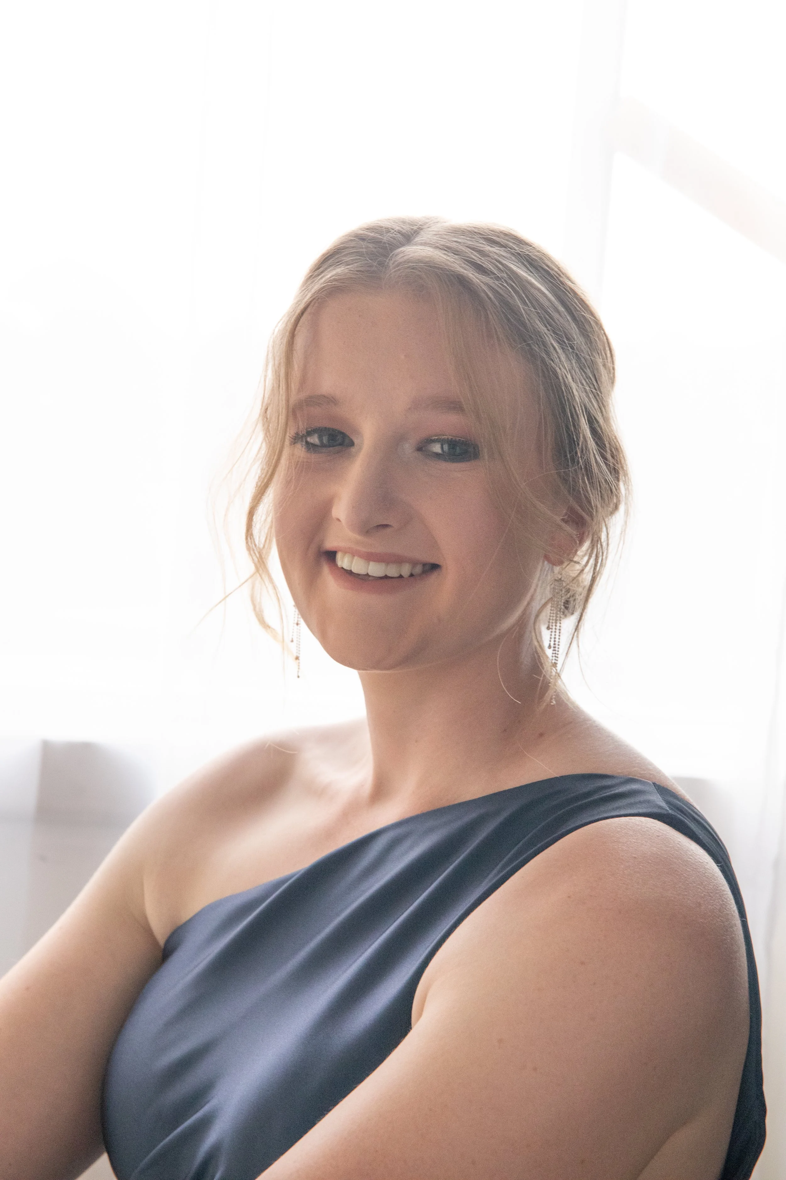 A smiling young woman with blonde hair in a loose updo, wearing a dark satin dress with one shoulder, standing in front of sheer white curtains.