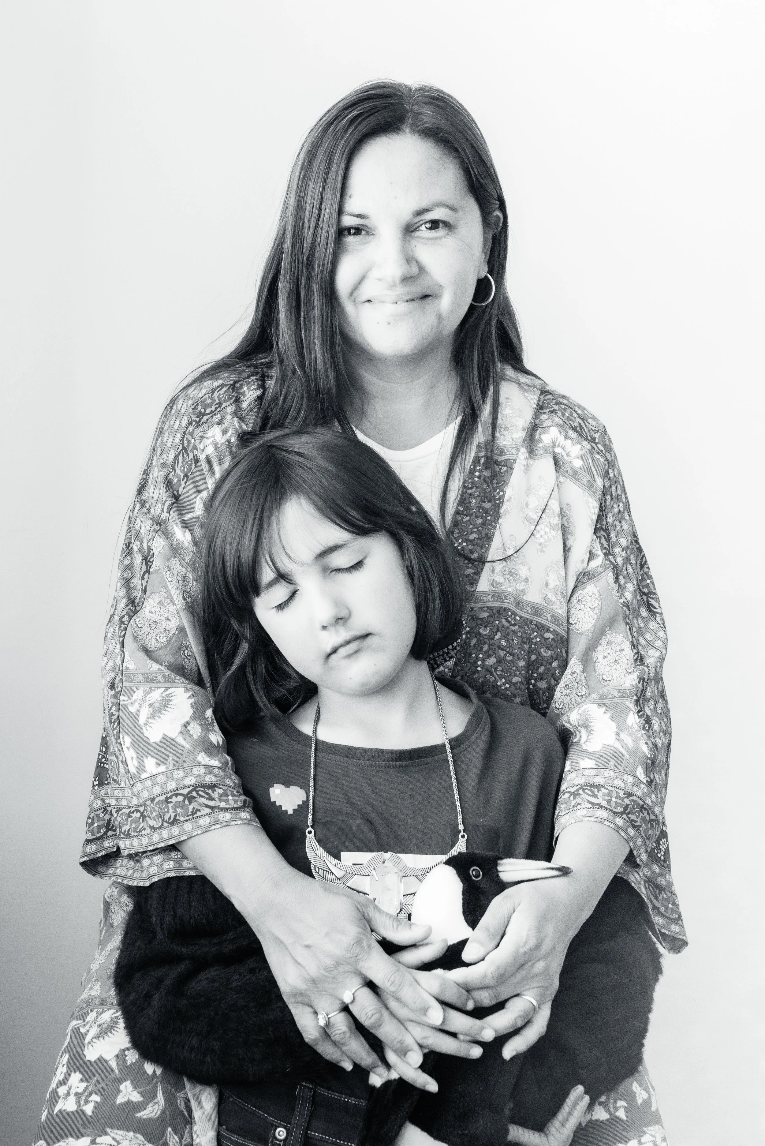 A woman and a young girl, both with dark hair, are posing for a black-and-white photo. The woman is smiling gently and standing behind the girl, who has her eyes closed and appears to be sleeping or resting. The woman is holding a plush penguin toy.