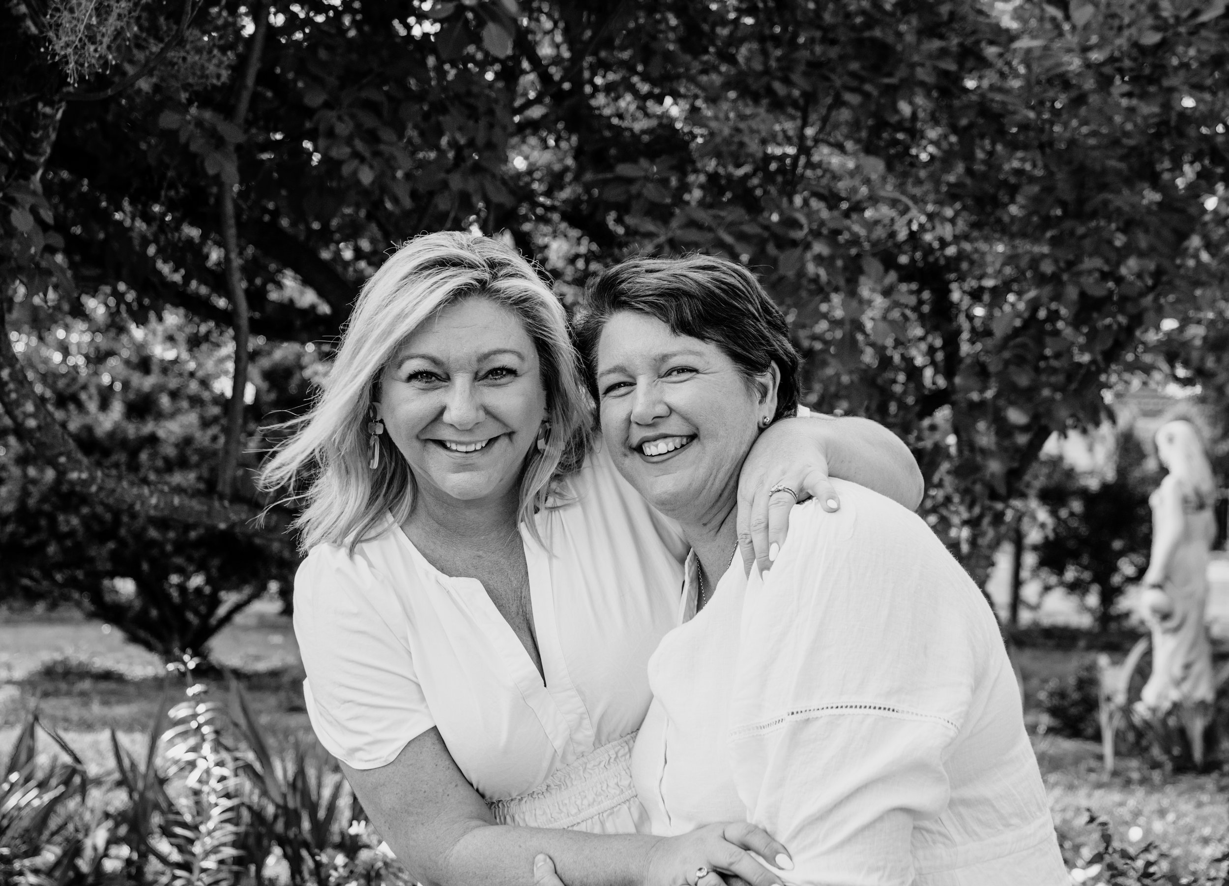 Black and white photo of two women smiling and embracing outdoors, with trees and sculptures in the background.