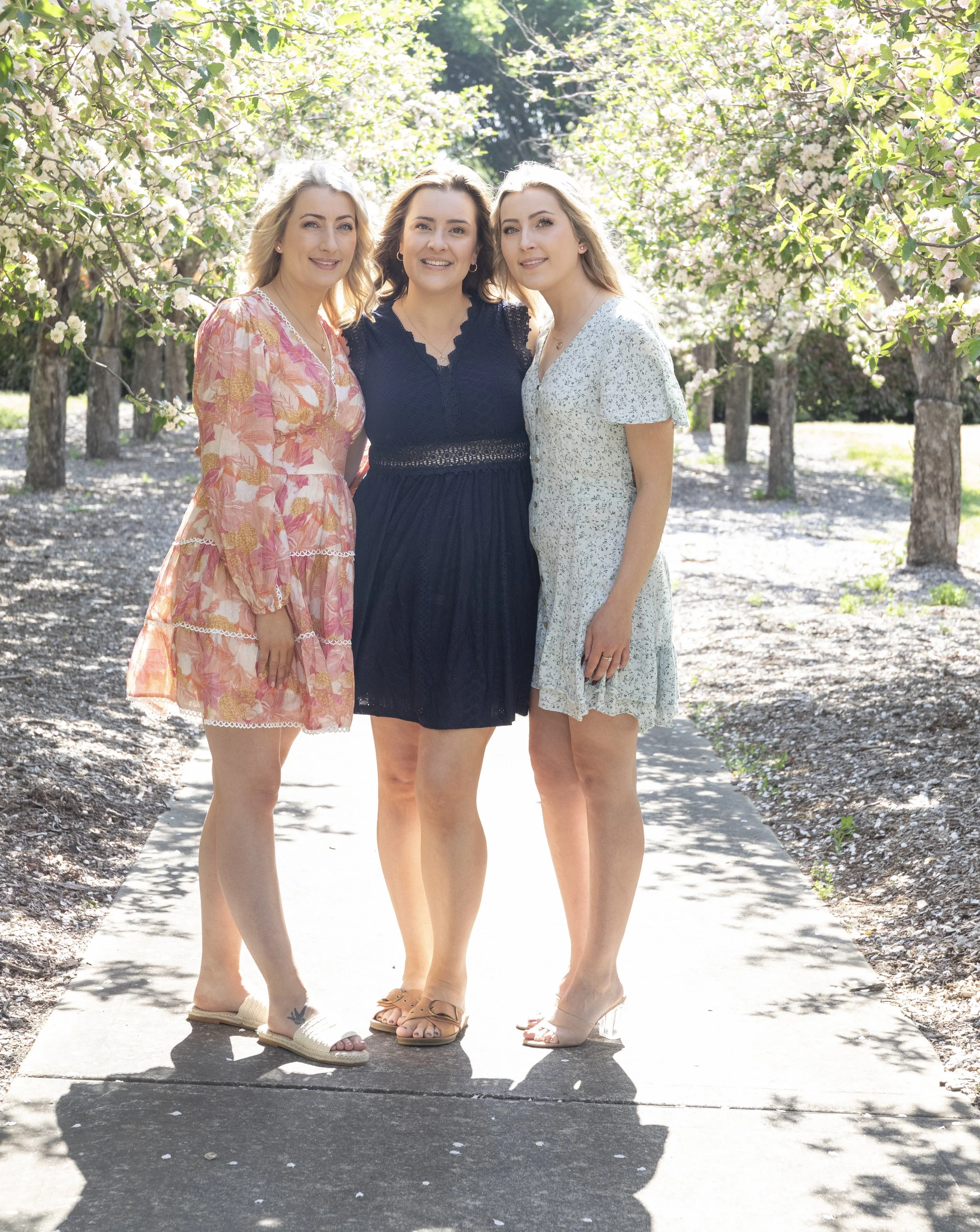 Three women standing together outdoors on a sunny day, surrounded by blooming trees.