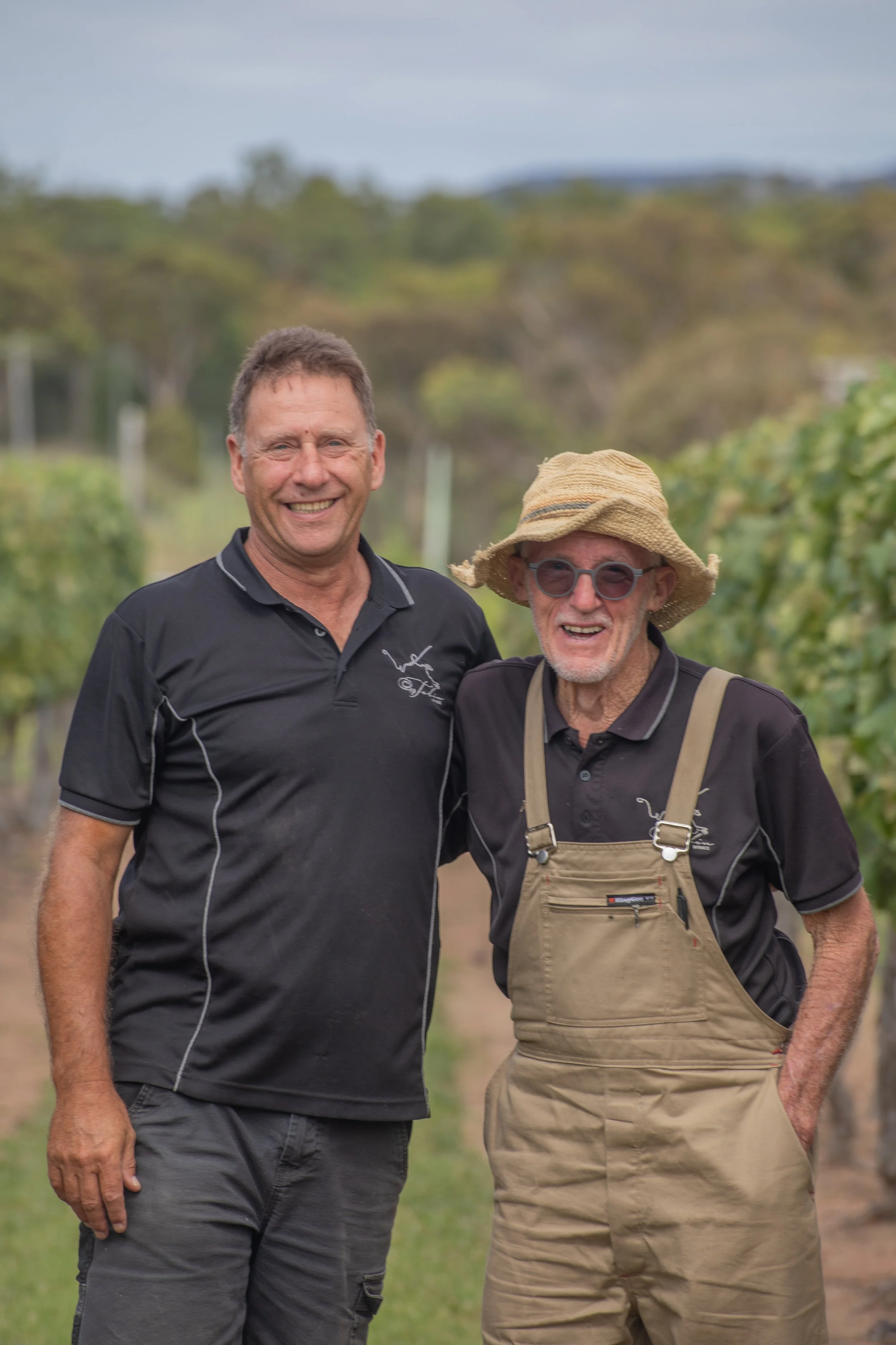 Two men standing together outside in a vineyard, smiling at the camera. One man is younger with short brown hair, wearing a black polo shirt and gray cargo pants. The older man has gray hair, glasses, a straw hat, and beige overalls over a black polo