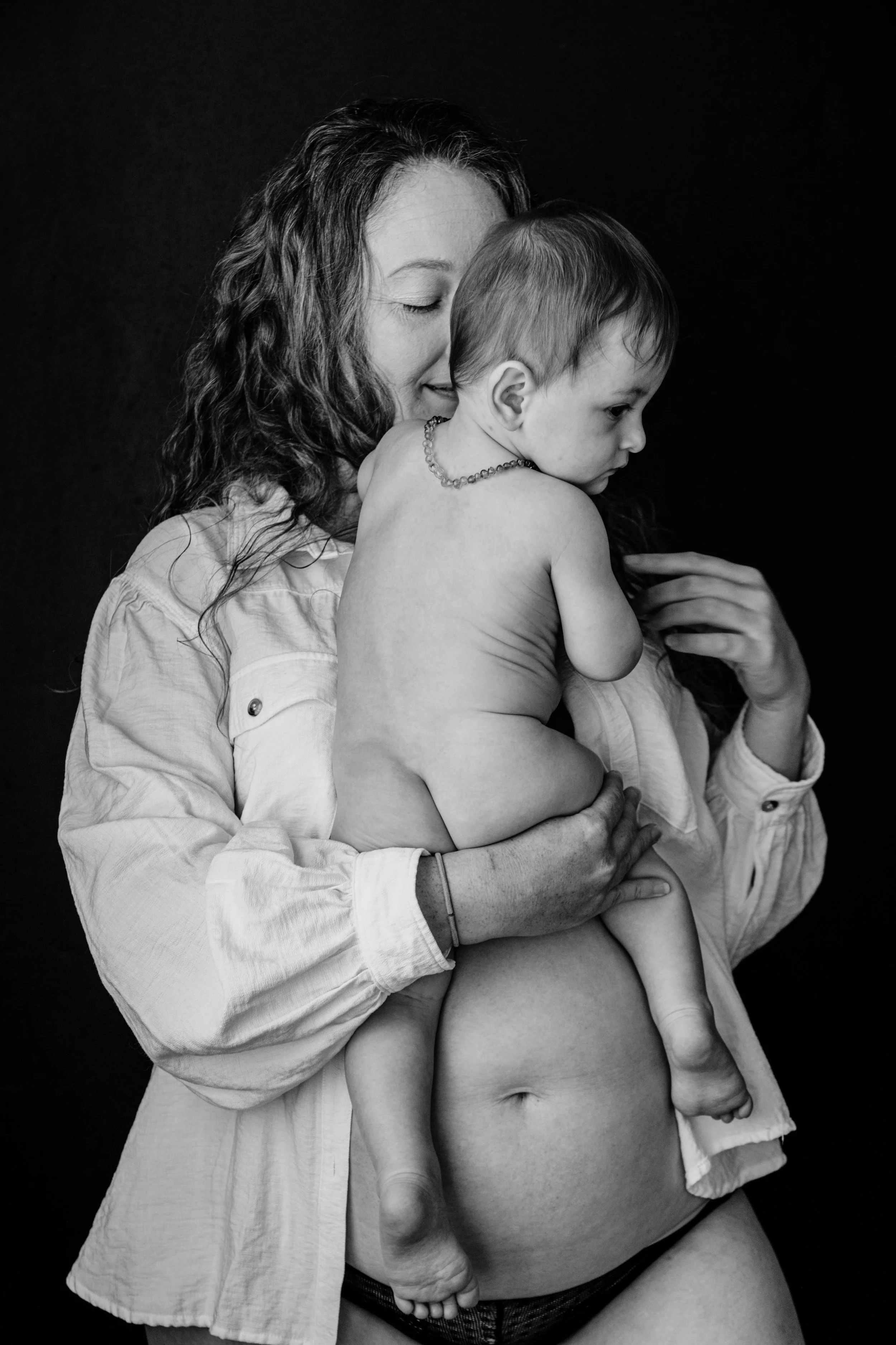 Black and white photo of a woman holding a naked toddler against a dark background.
