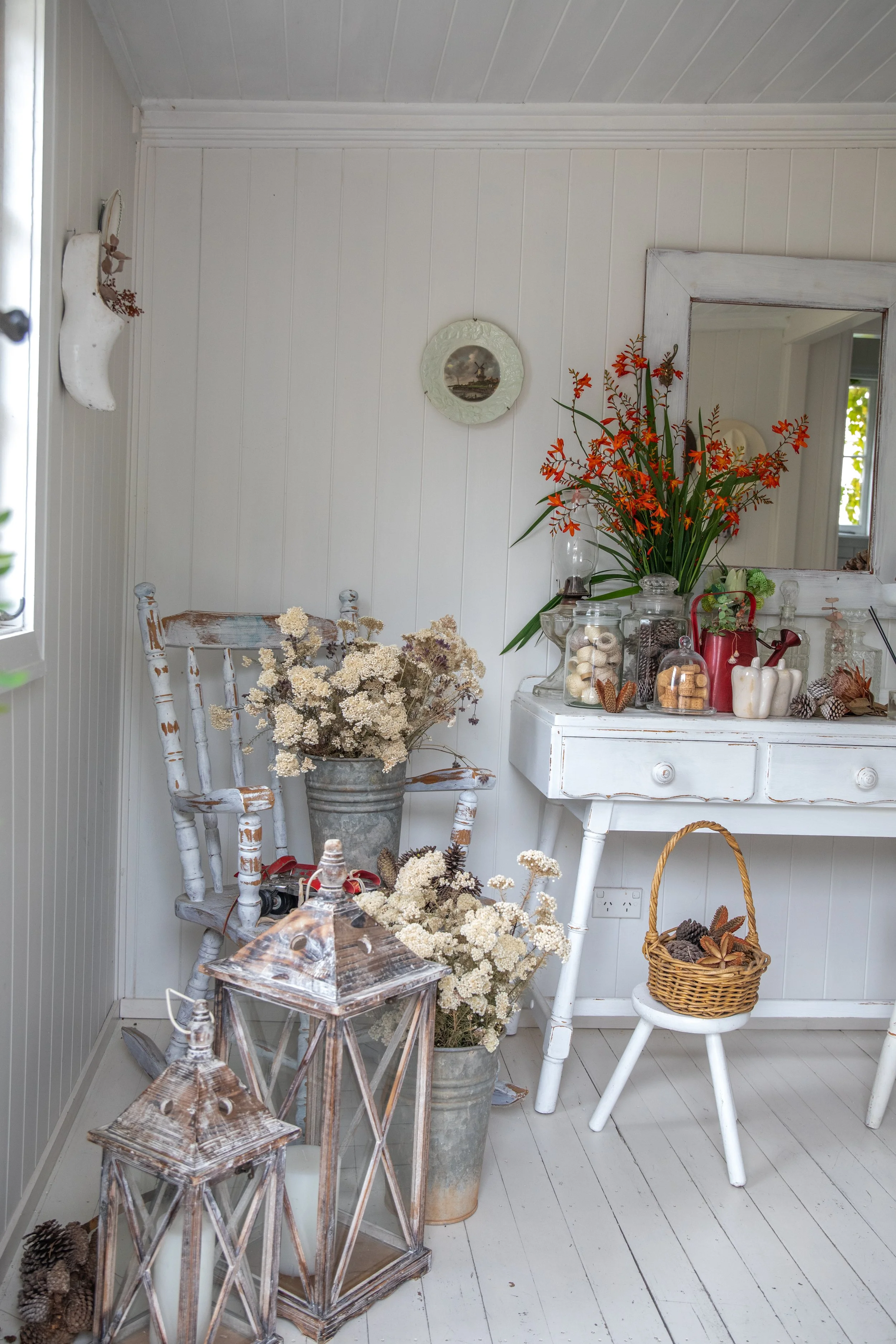 A cozy nook with white painted wooden walls and floors, decorated with vintage white furniture and floral accents. There is a white dresser topped with glass jars, flowers, and autumnal decorations. A white chair with a basket of pinecones and leaves