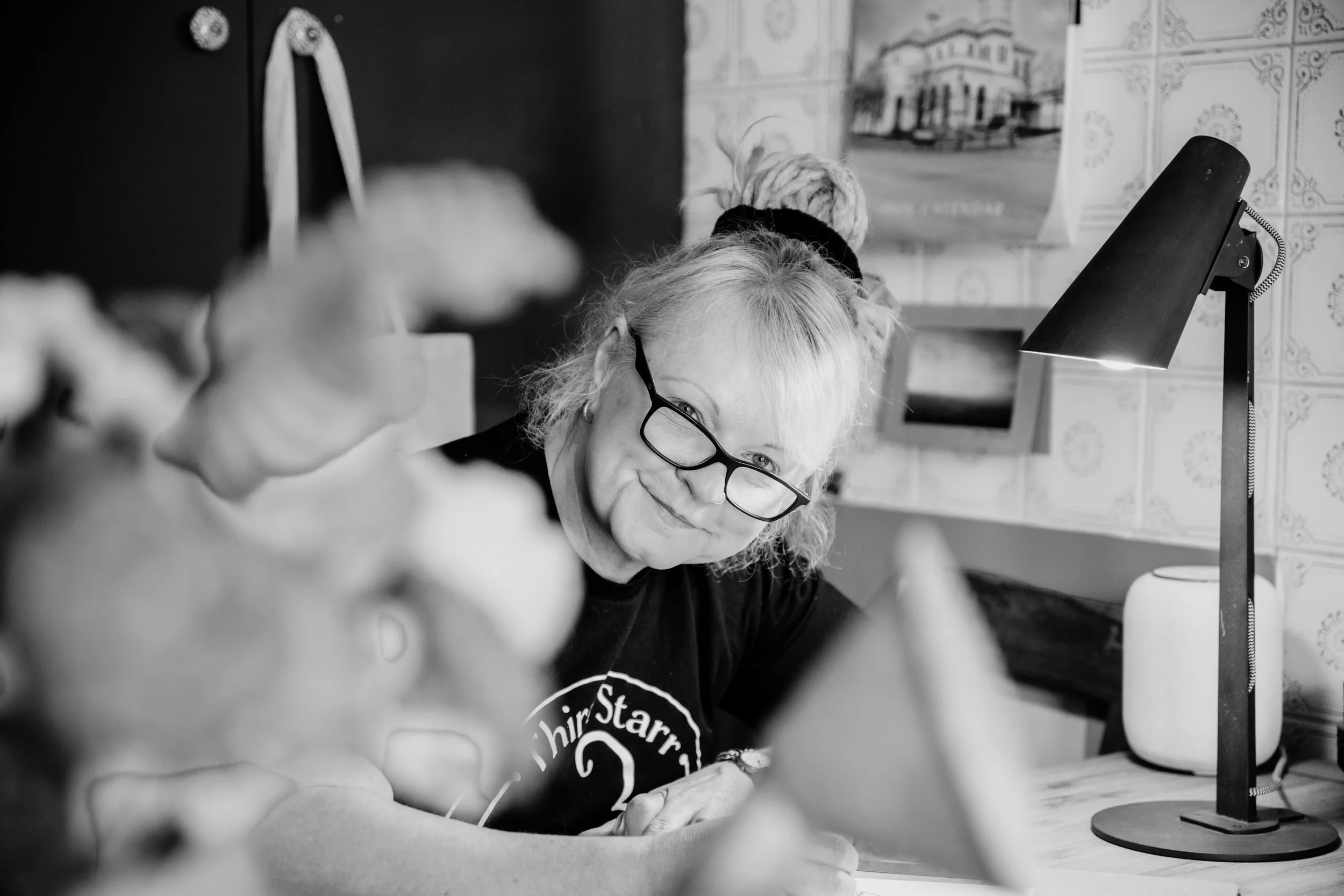 A woman with glasses and blonde hair in a bun, smiling while working at a desk with a lamp and blurred objects in the foreground.