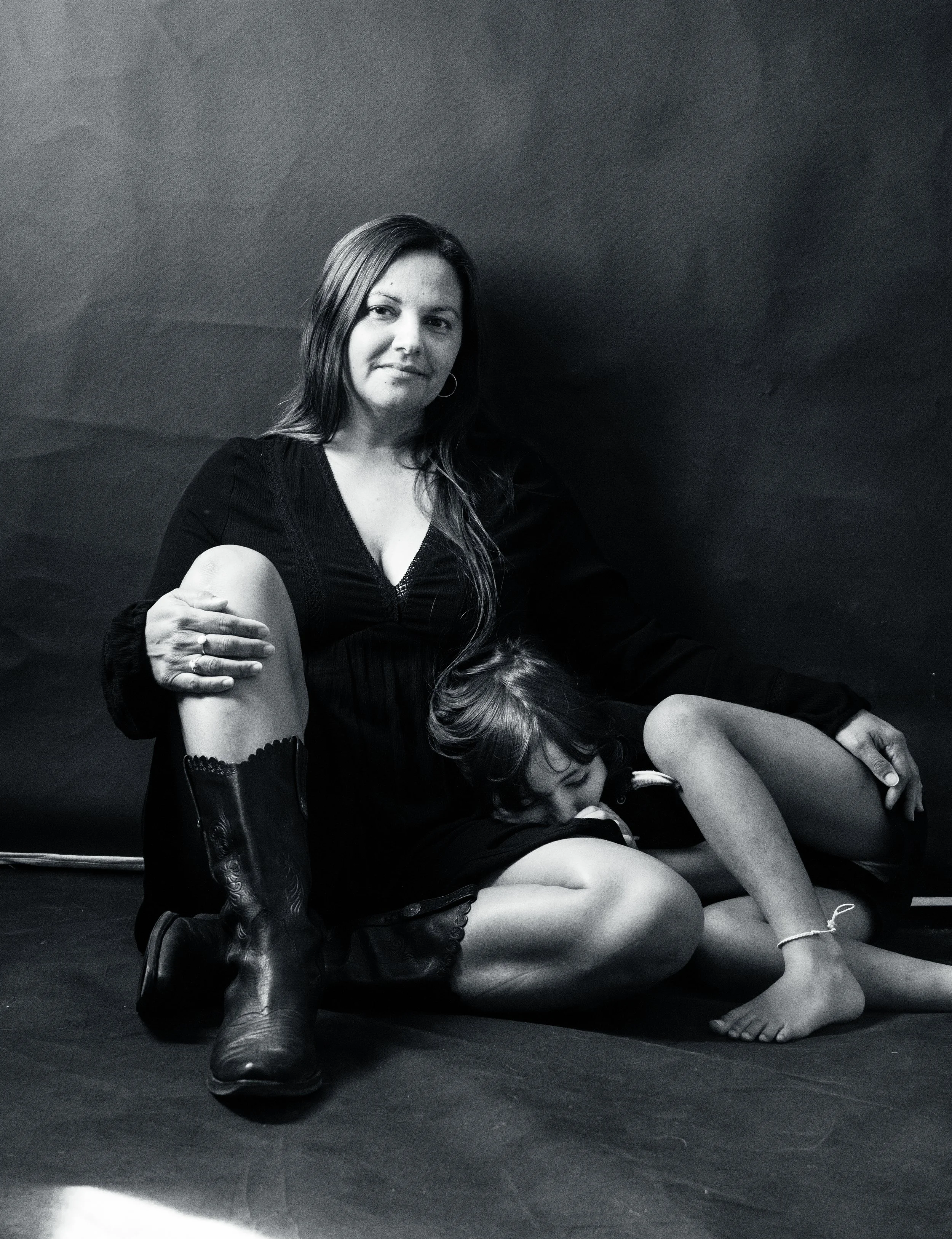 A woman sitting on the floor with a young girl resting her head on her lap. The woman has medium-length hair, wearing a black dress, and the girl has short hair, barefoot, with an ankle bracelet. The scene is in black and white.