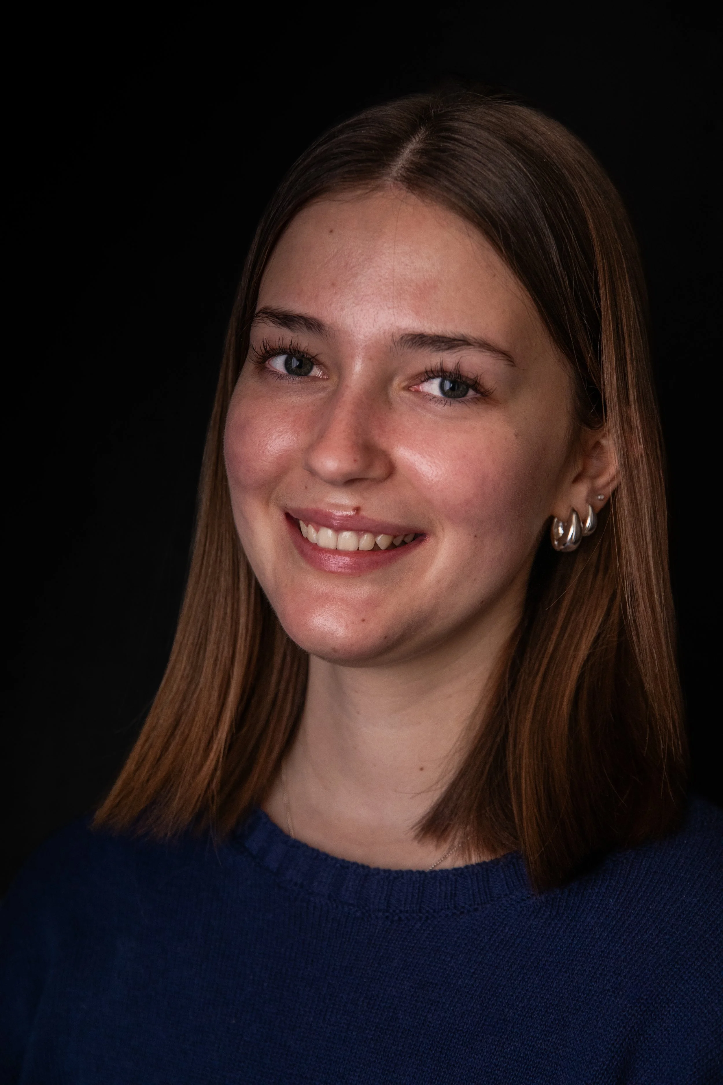 A young woman with light skin, shoulder-length brown hair, blue eyes, and silver hoop earrings, smiling against a black background, wearing a navy blue top.