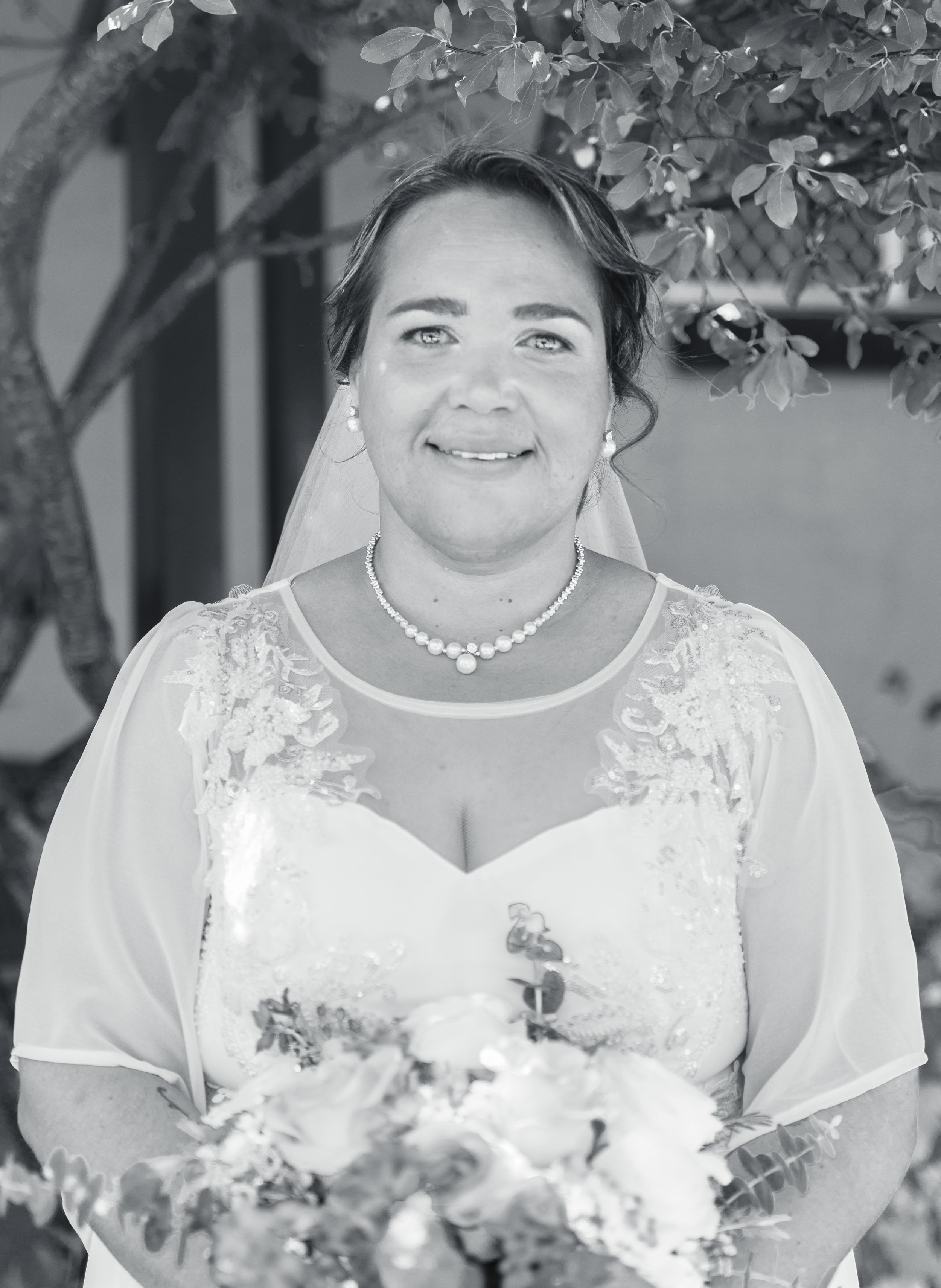 A woman in a wedding dress holding a bouquet, standing outdoors in front of a tree and building.