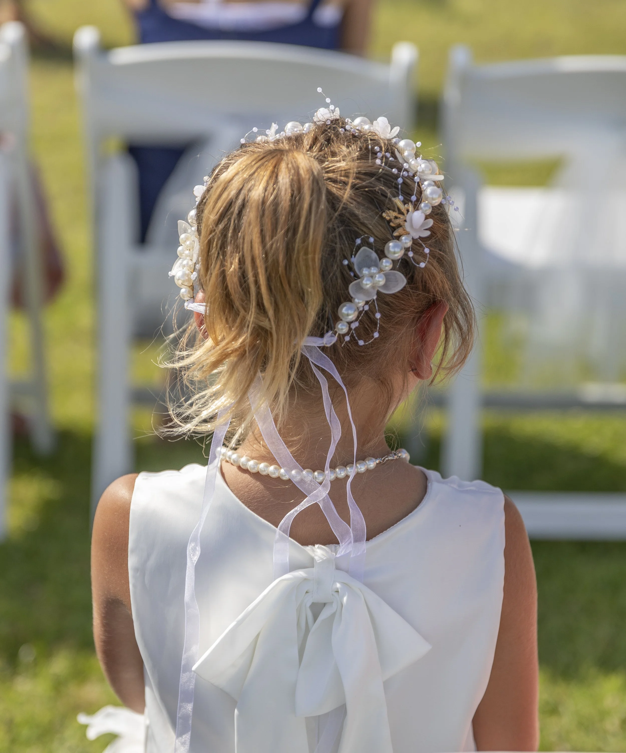 Back view of a young girl in a white dress with a bow, wearing a pearl necklace, with her hair styled in a ponytail adorned with a pearl and flower headband, at an outdoor event with white chairs.