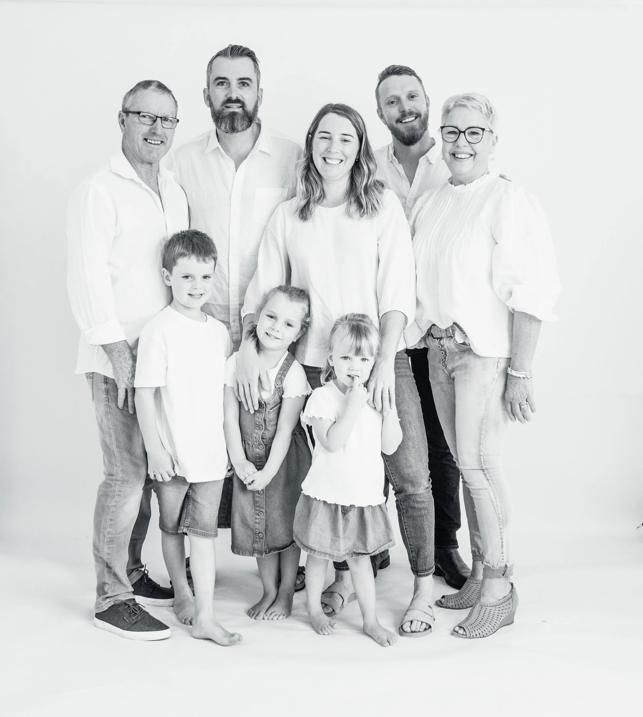 Black and white photo of a multi-generational family group smiling and posing together indoors against a white background. The group includes adults and children, all casually dressed.
