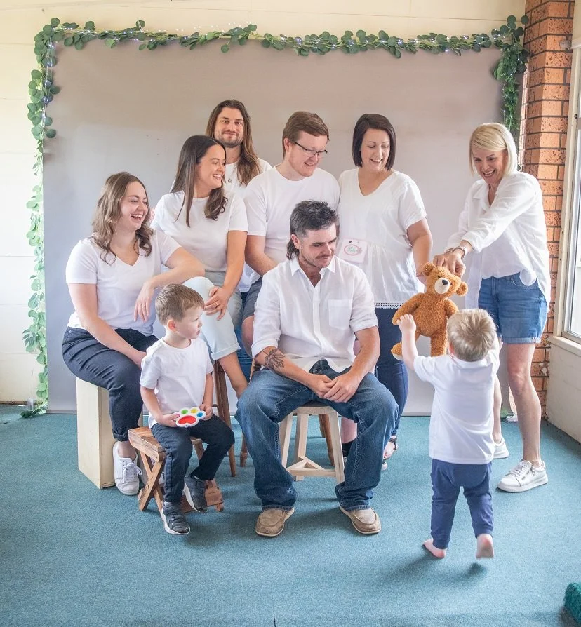 A group of adults and children gathered for a photo, with some sitting and others standing, as a woman hands a teddy bear to a young boy in front of a backdrop decorated with green garland.