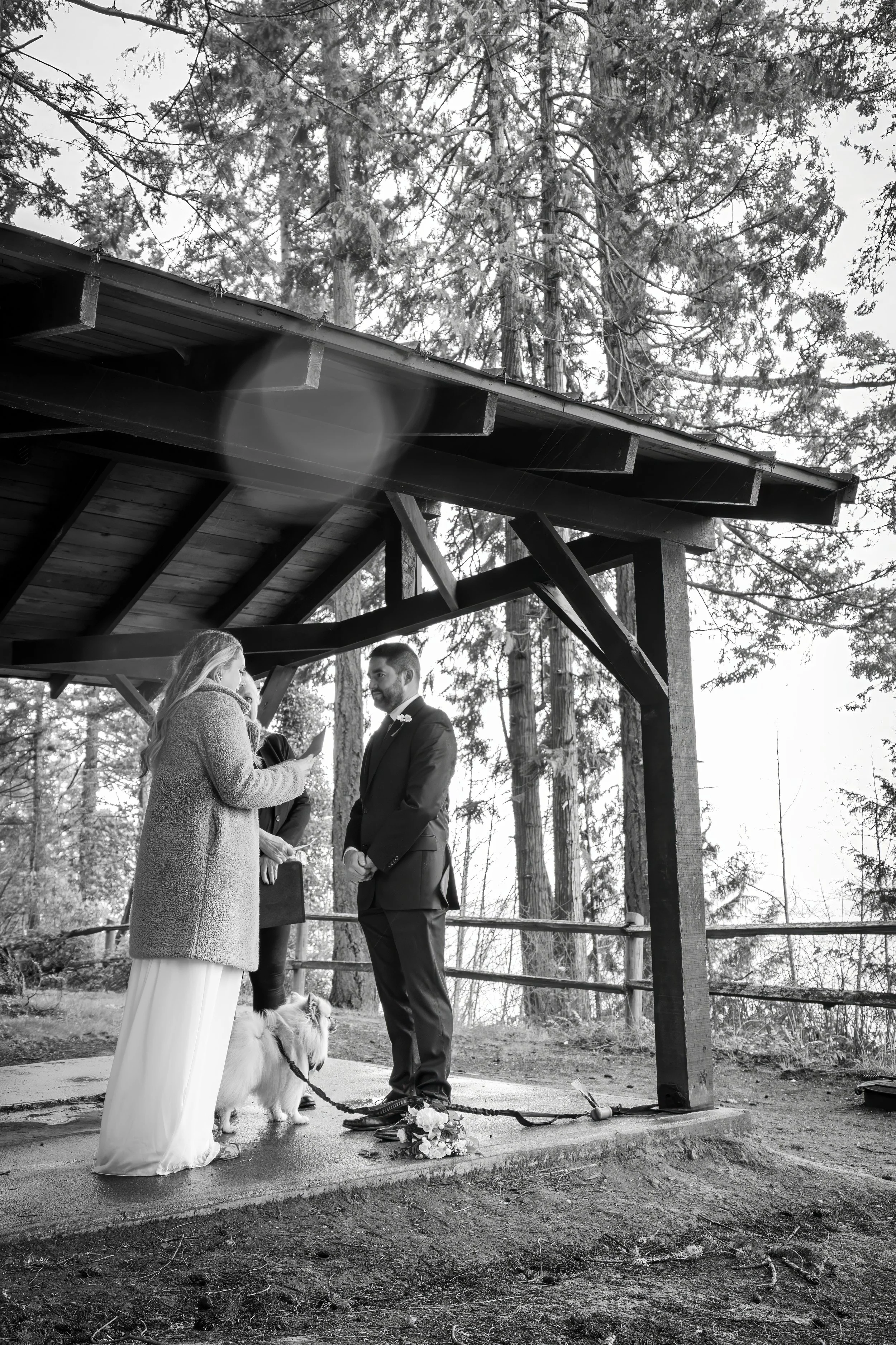 A black and white photo of a wedding ceremony outdoors under a wooden pavilion, with a bride and groom standing in front of each other. The bride is speaking, holding a phone, and the groom is listening. A woman is standing behind them, and a dog is 