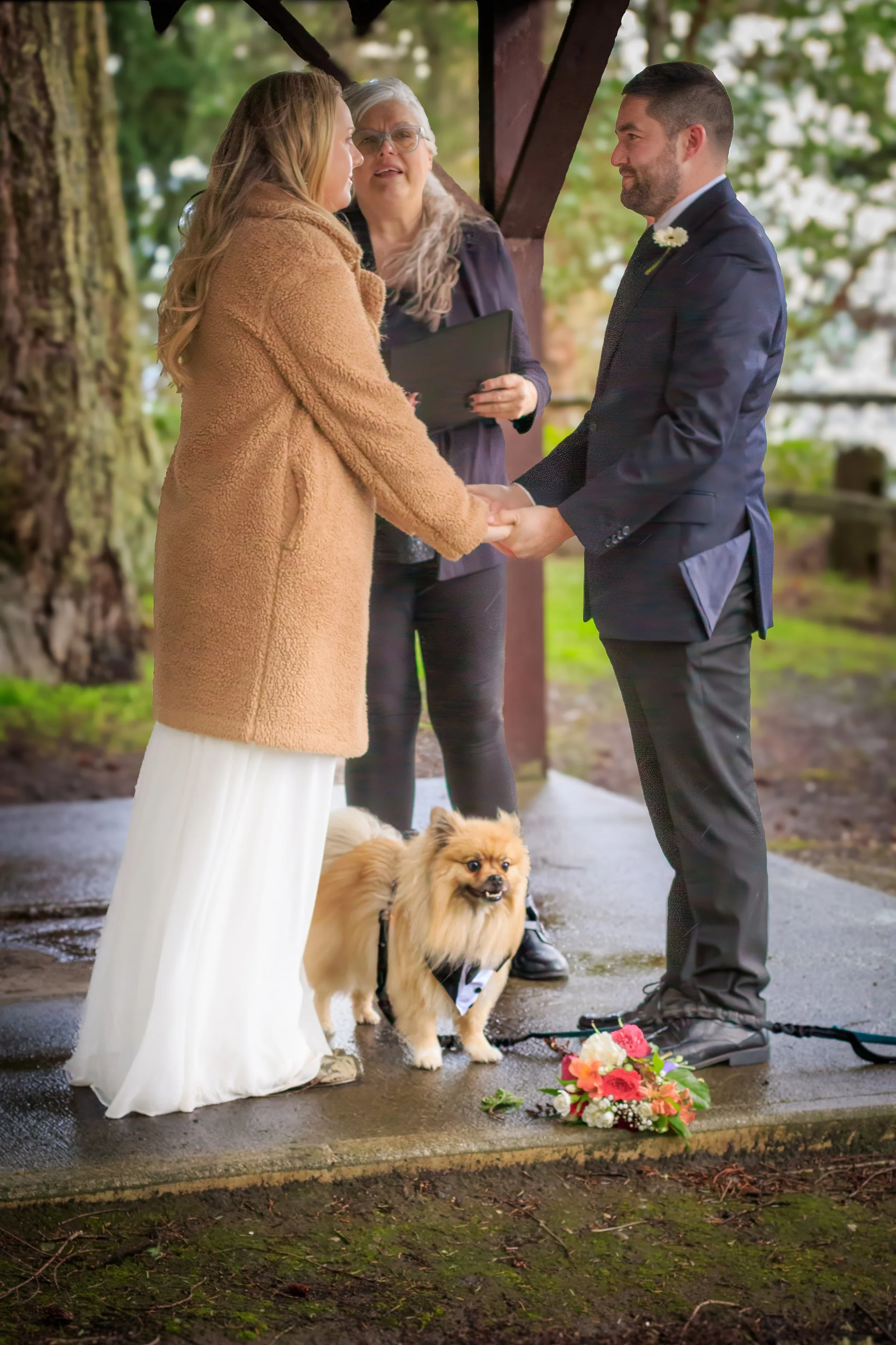 A couple is getting married outdoors under a wooden structure, holding hands, with a officiant standing behind them. The woman is wearing a white dress and a tan coat, and the man is in a dark suit with a white flower boutonniere. A small dog on a ha