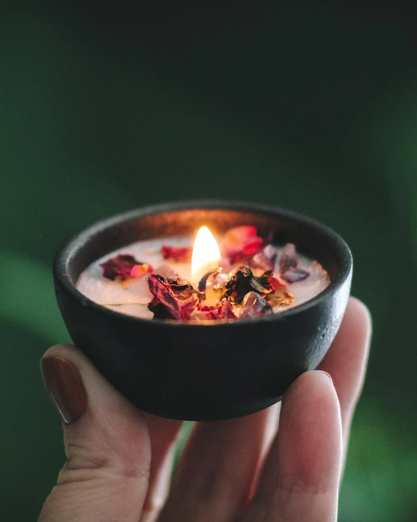A hand holding a small black bowl with a lit candle surrounded by dried flower petals and crystals.