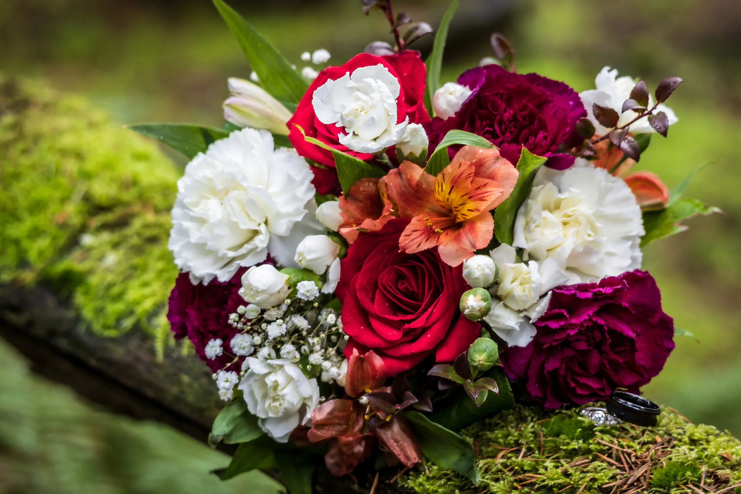 A colorful bouquet of flowers including red roses, white carnations, orange lilies, and purple carnations, resting on moss-covered wood in a natural outdoor setting.