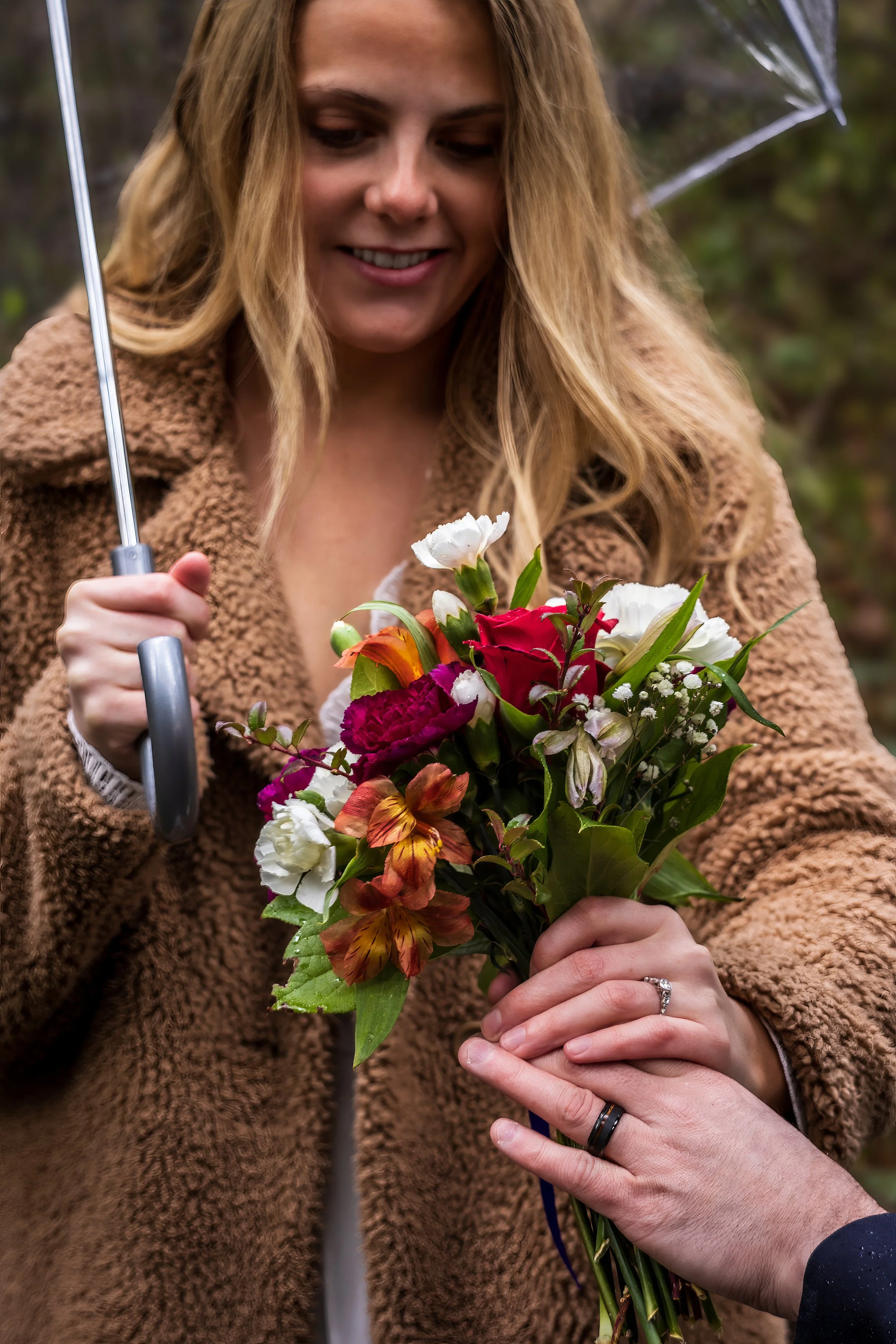A woman holding a bouquet of colorful flowers with a man’s hand handing it to her outdoors, with an umbrella and blurred background.