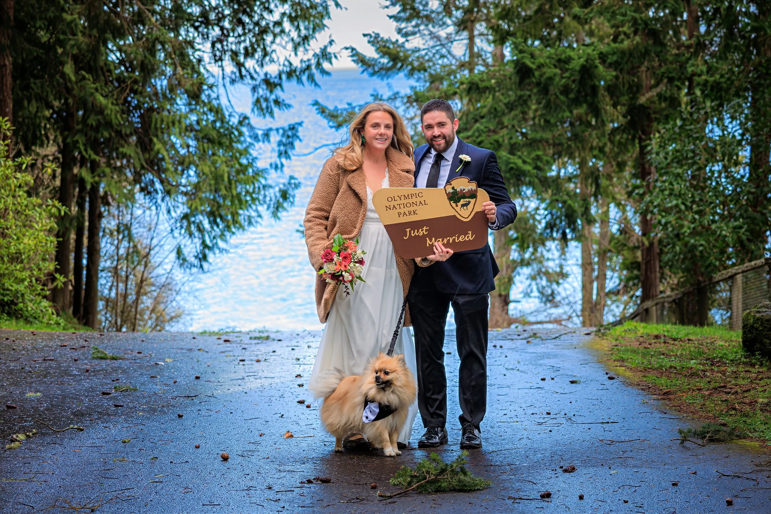 A newly married couple outdoors near a lake, holding a sign that reads 'Olympic National Park, Just Married,' with a woman in a white dress holding a bouquet and a man in a suit. They are accompanied by a small fluffy dog wearing a tuxedo. The backgr