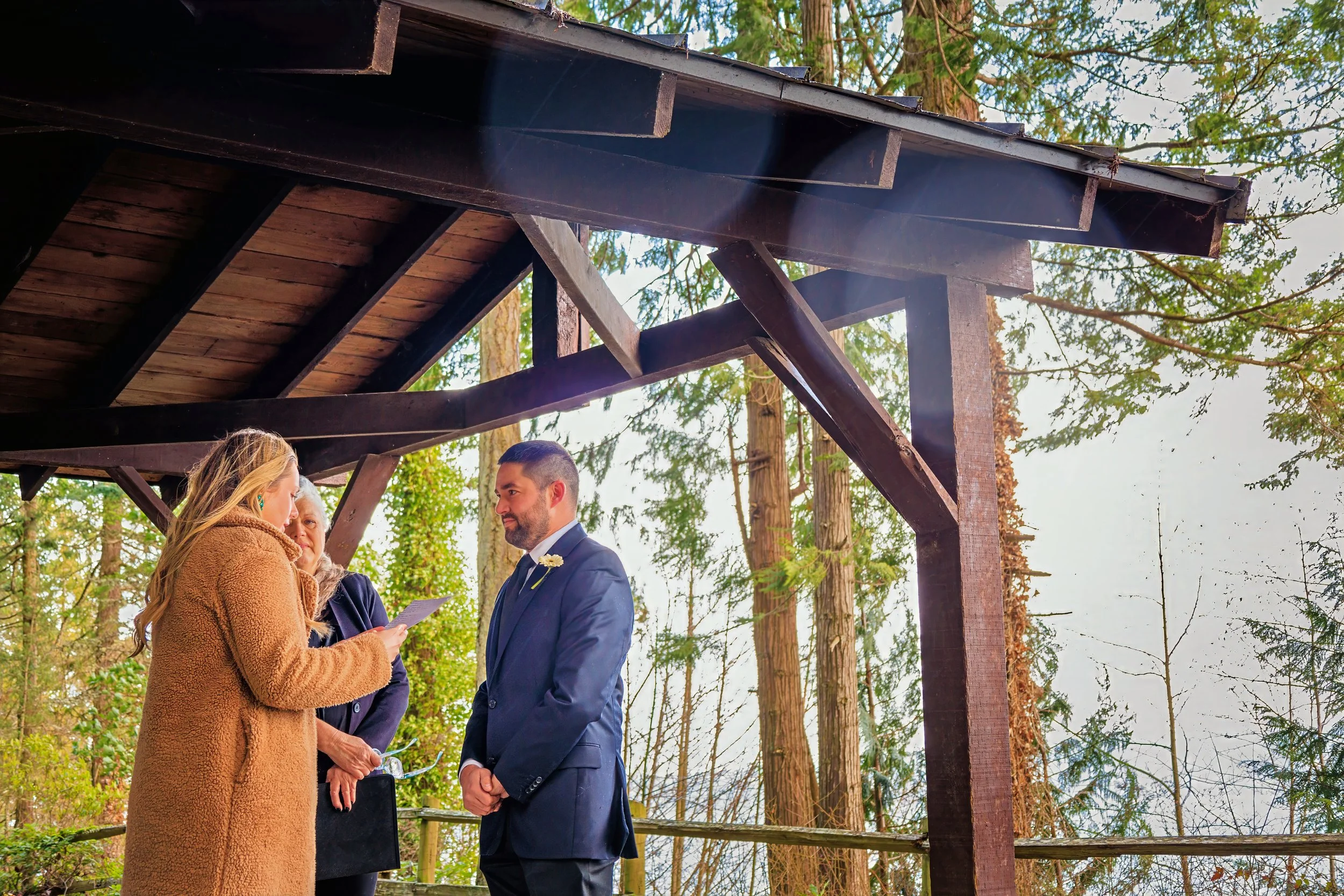 A wedding ceremony outdoors under a wooden pavilion in a forest with three people, including a man in a blue suit and tie and two women, one in a tan coat and the other in a dark blazer, standing on a wooden deck.