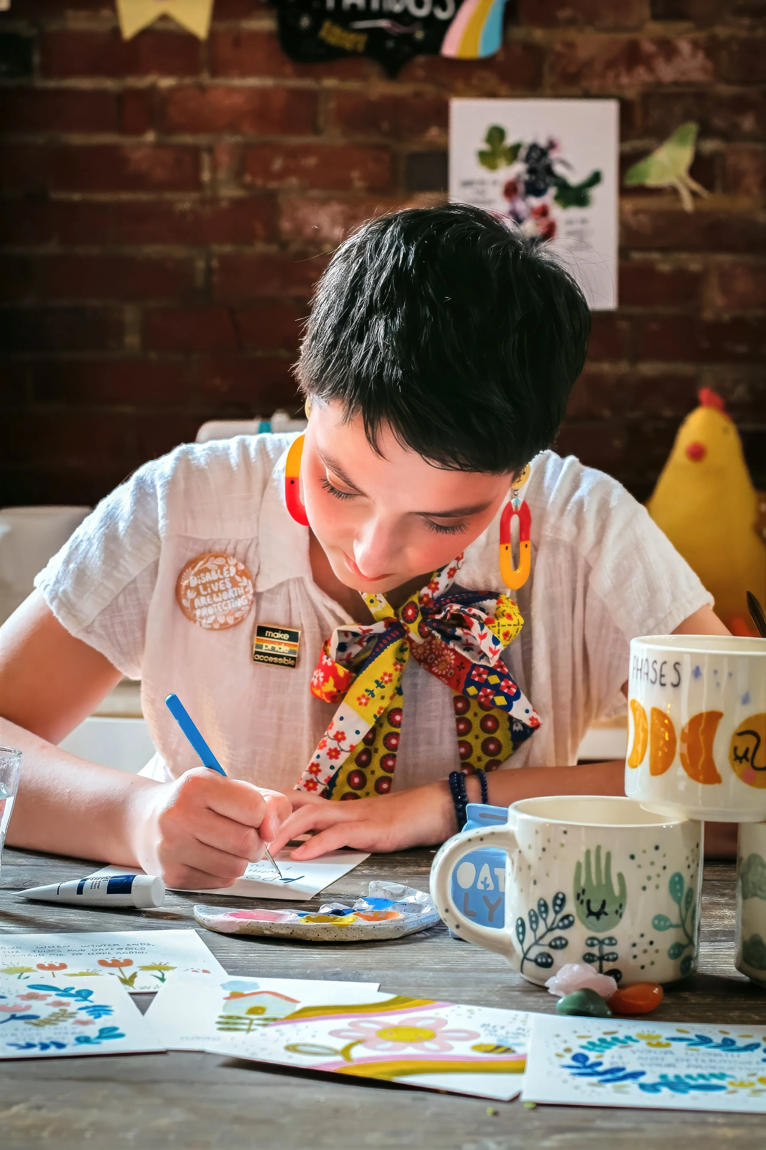 A person with short dark hair drawing or writing on paper at a table with cups and art supplies, in a room with a brick wall background.