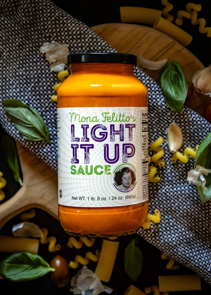 A jar of Mona Felitto's Light It Up Sauce placed on a wooden cutting board, surrounded by scattered garlic, basil leaves, and pasta shapes on a black and white patterned cloth.