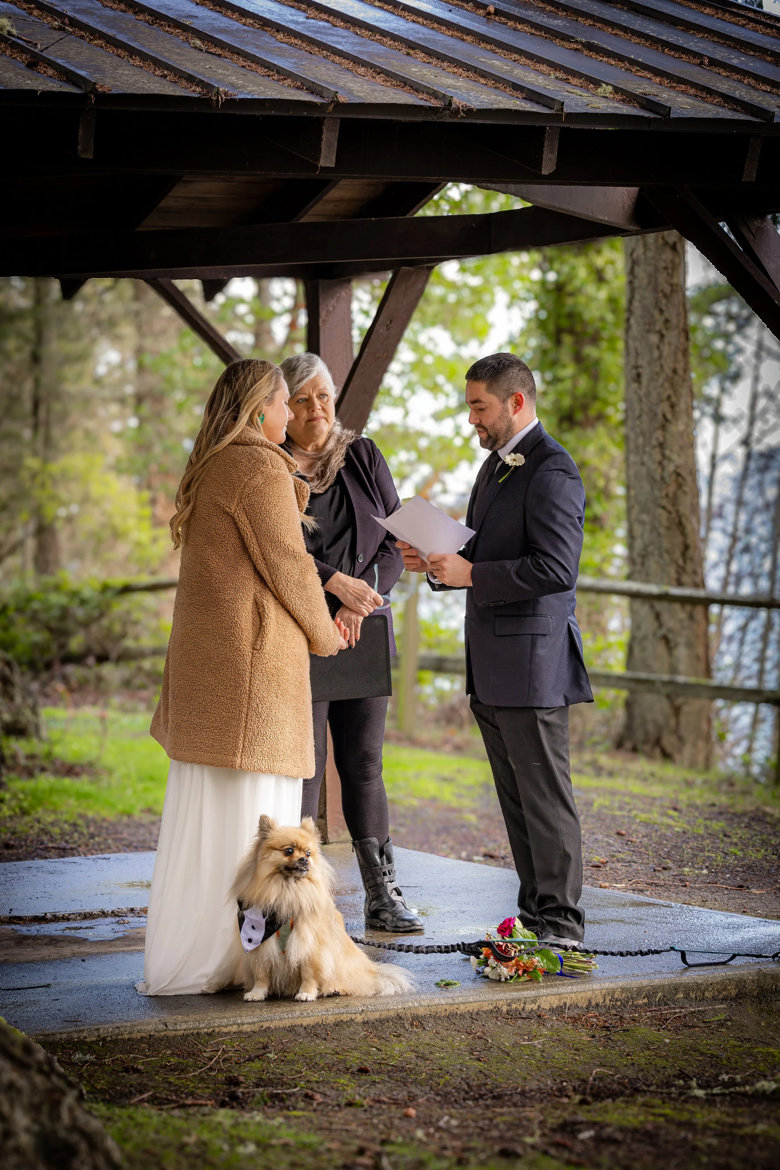 A wedding ceremony taking place outdoors beneath a wooden structure, with a couple holding hands and an officiant reading from a paper. A small fluffy dog with a bow tie and leash sits in front of the bride, with a bouquet of flowers on the ground ne