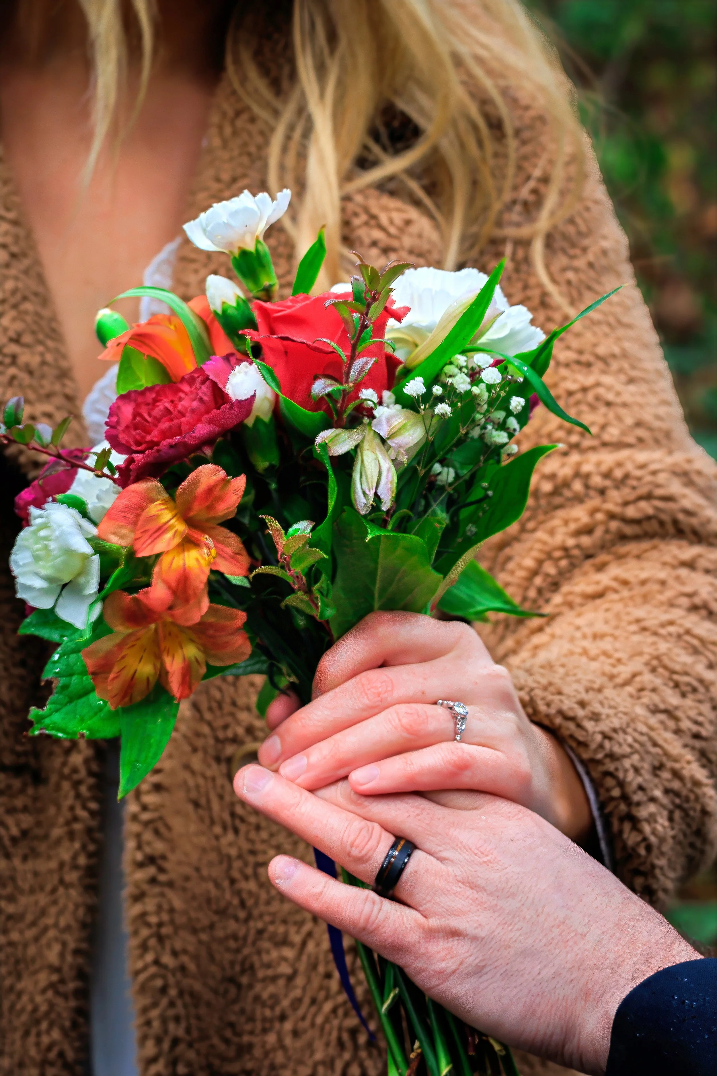 A woman in a brown, fluffy sweater is holding a vibrant bouquet of mixed flowers, including roses and lilies, with another person’s hand gently touching the bouquet; the person has a wedding or engagement ring on their finger.