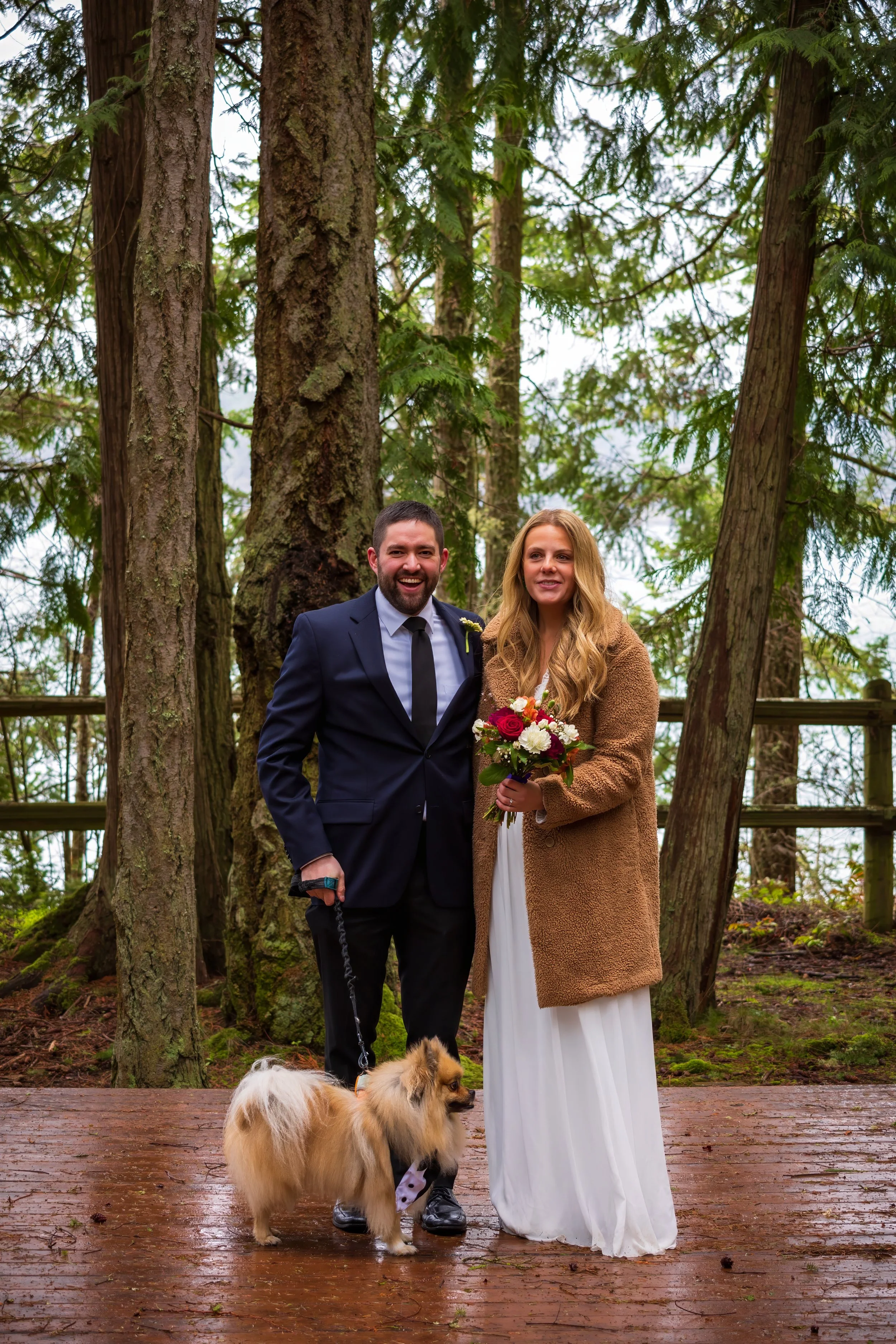 A couple dressed for a wedding, standing outdoors on a wooden platform in a forest, with a small dog on a leash, posing for a photo.