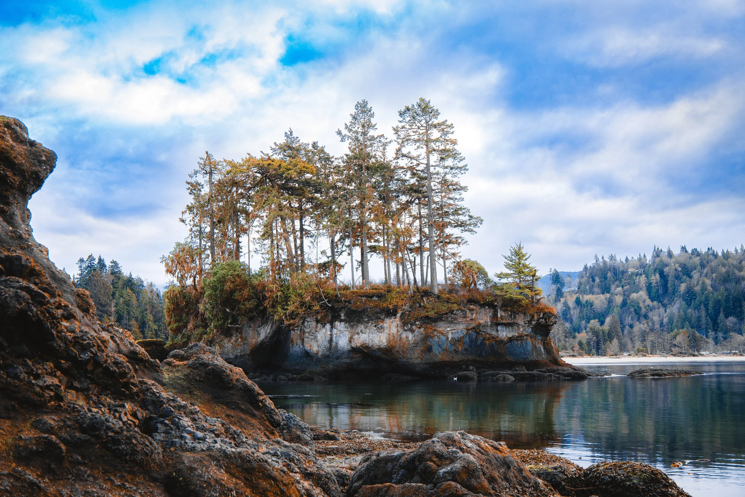 A rocky shoreline with a small island covered in trees, reflected in calm water, under a partly cloudy sky.