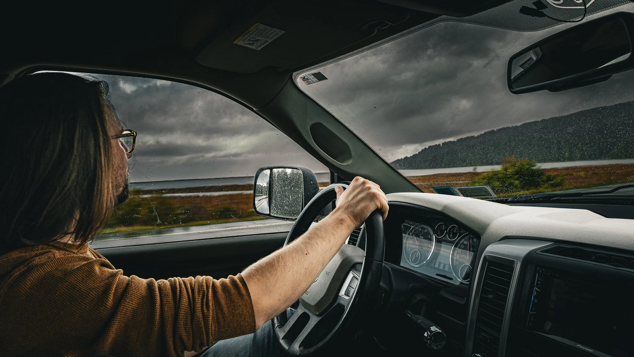 A person with long hair and glasses driving a vehicle during a rainy day with dark clouds and a landscape of fields and hills outside.