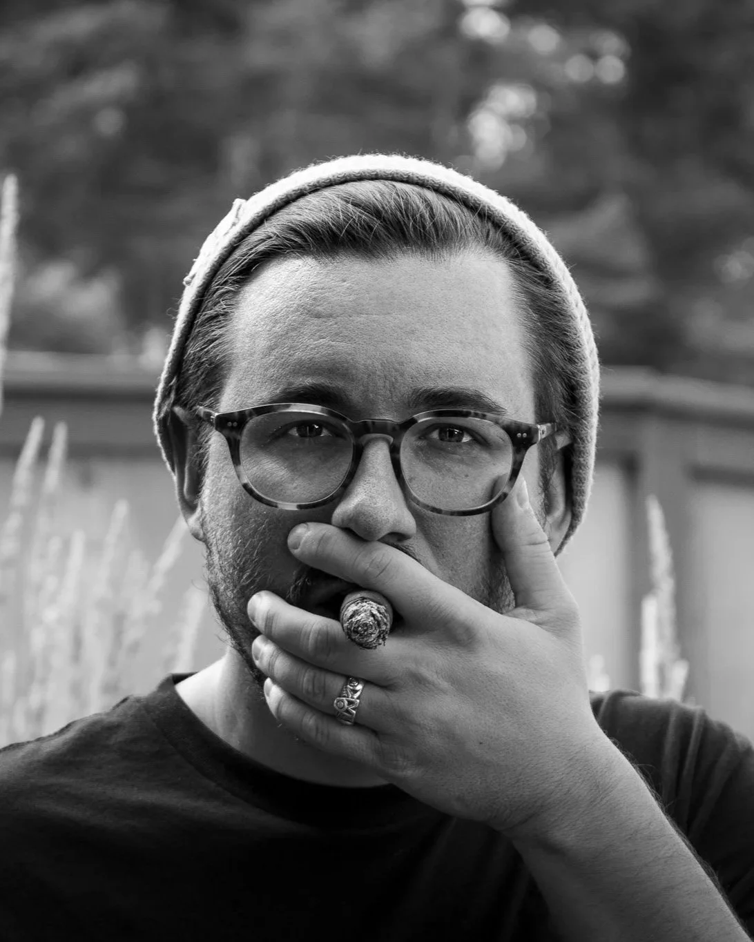A man with glasses and a beanie, holding a cigar in his mouth, outdoors.