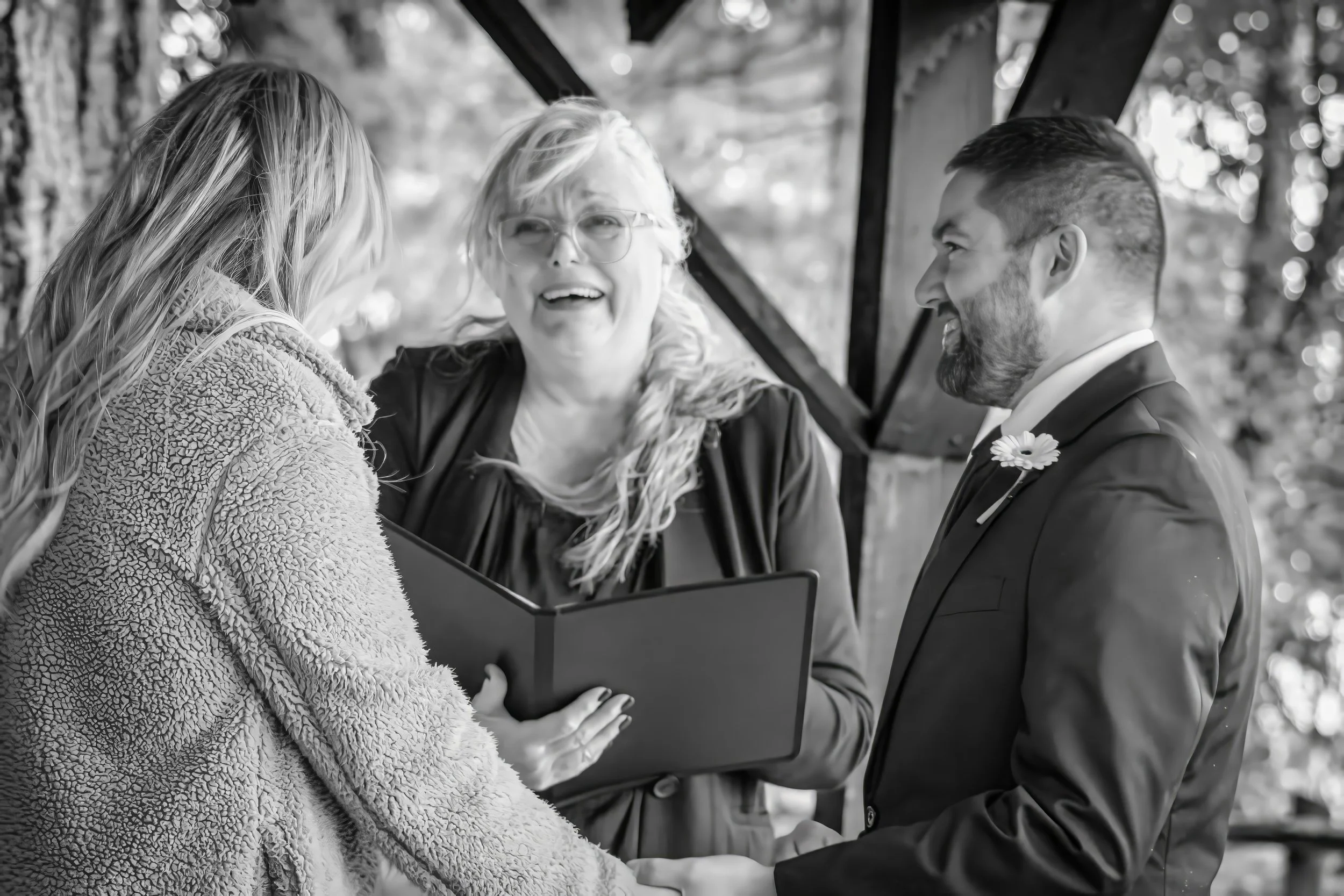 A couple getting married, holding hands and smiling at each other while a woman officiant reads from a book during an outdoor ceremony.