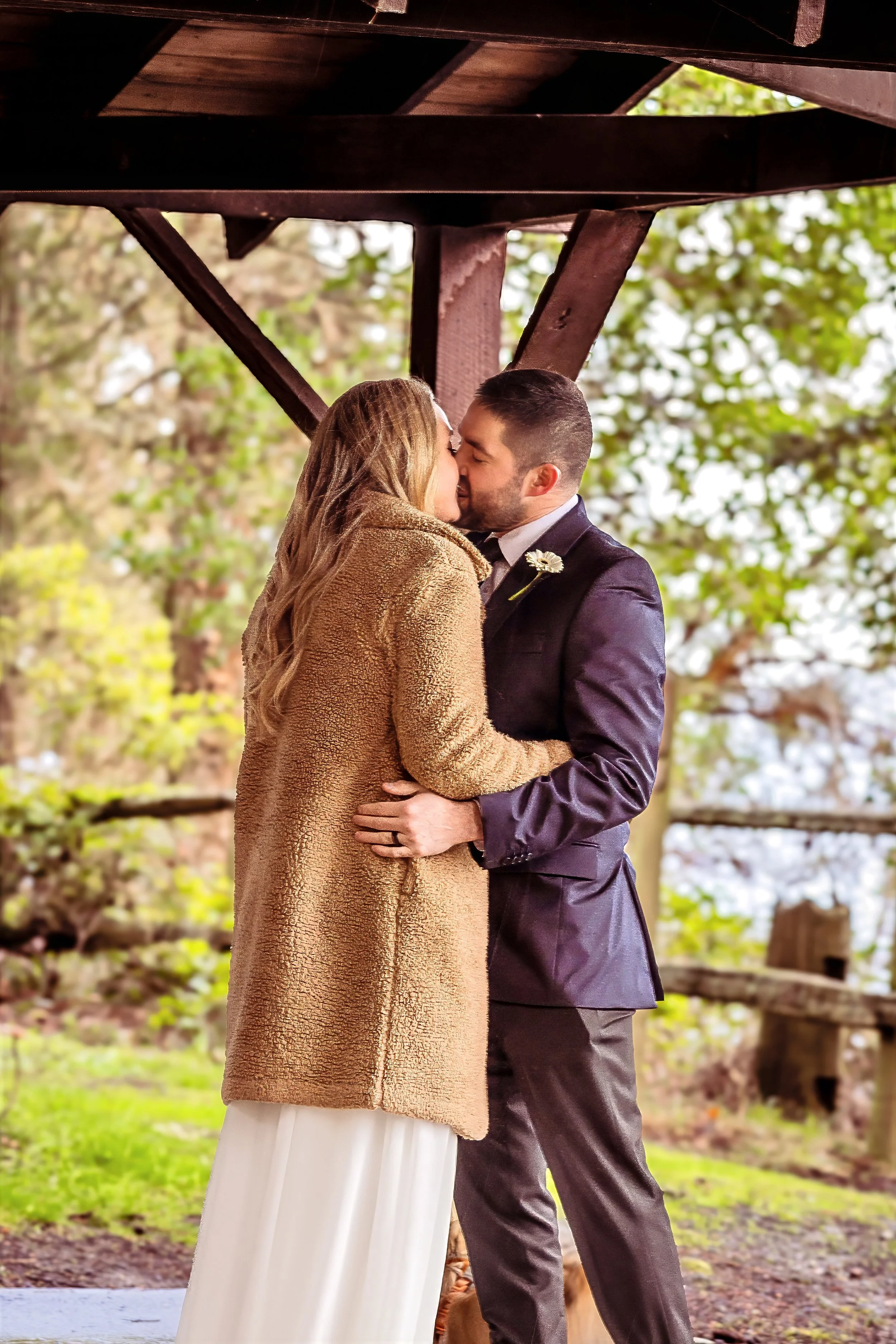 A couple is sharing a kiss outdoors under a wooden structure in a natural setting with trees and greenery.