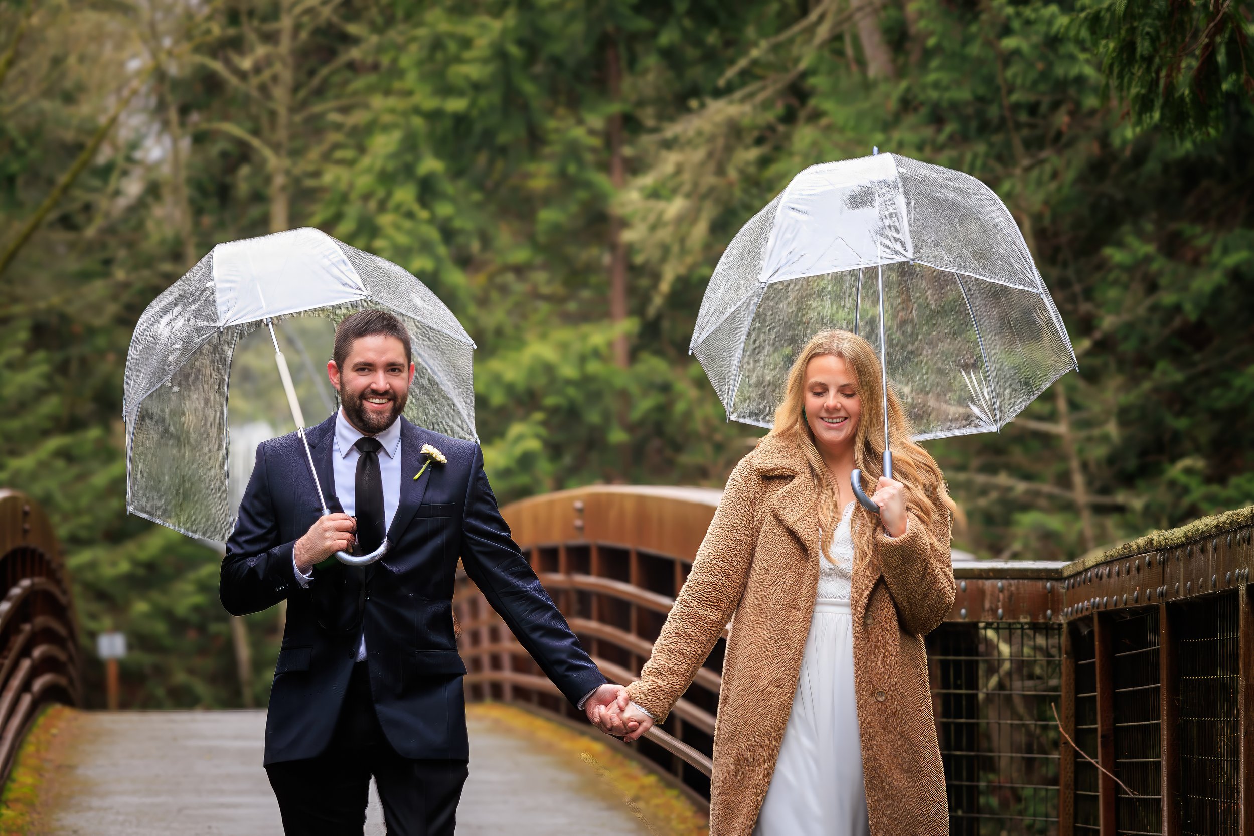A smiling couple holding hands and walking with transparent umbrellas on a wooden bridge in a green forested area.