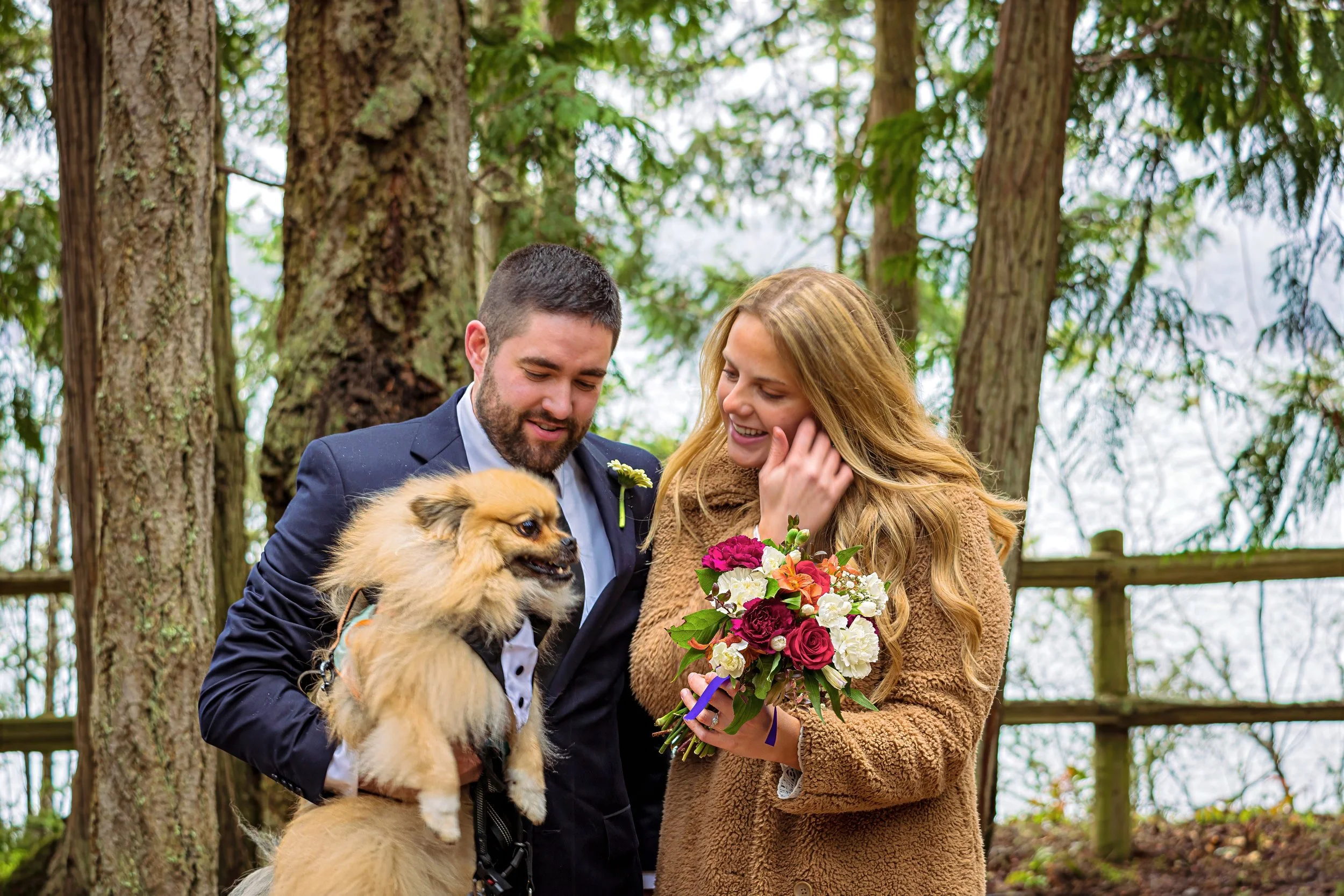 A couple dressed in wedding attire, standing outdoors among trees, with a woman holding a bouquet of flowers and a man holding a small dog, smiling and enjoying the moment.