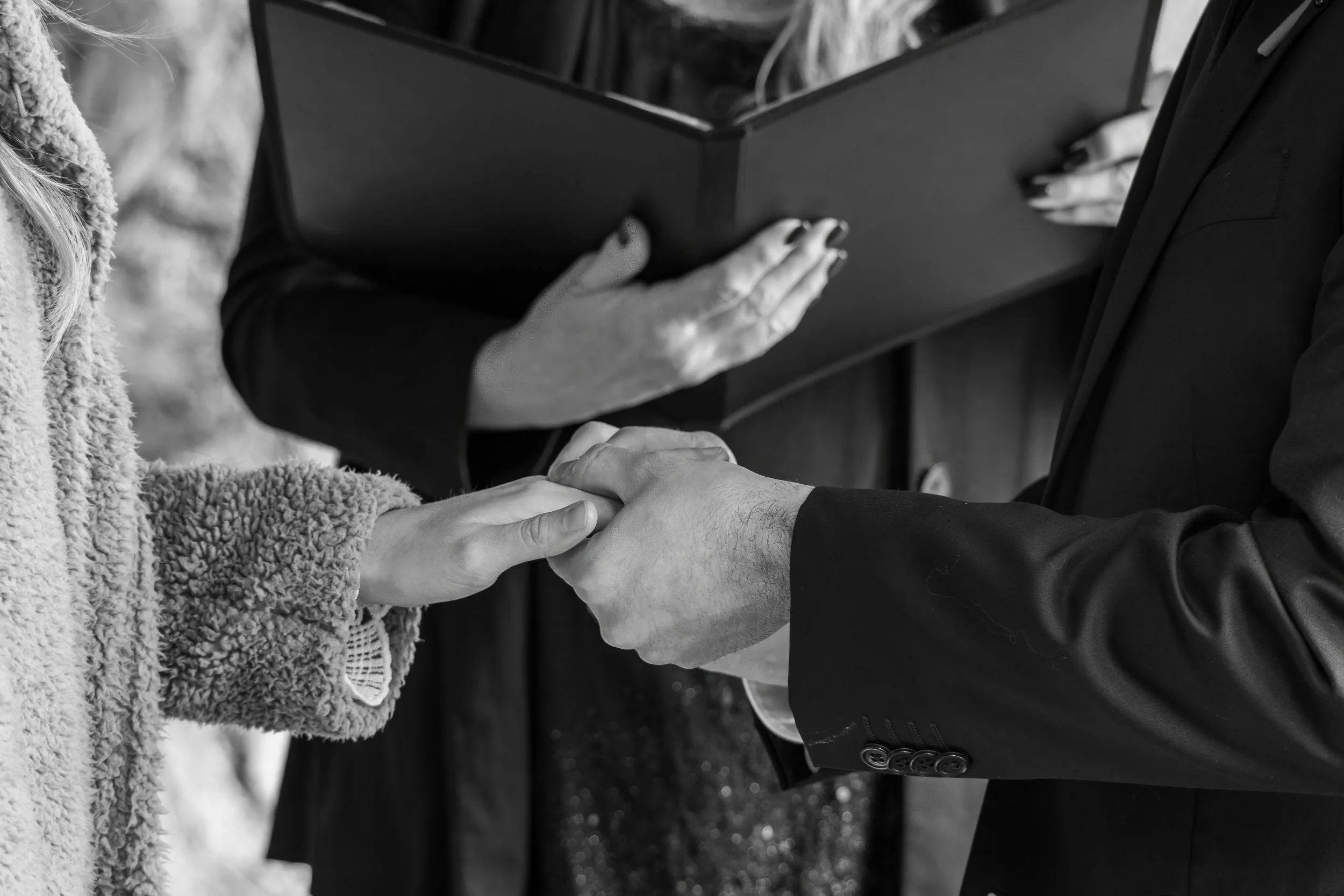 A couple holding hands during a wedding ceremony, with an officiant reading from a book in the background.