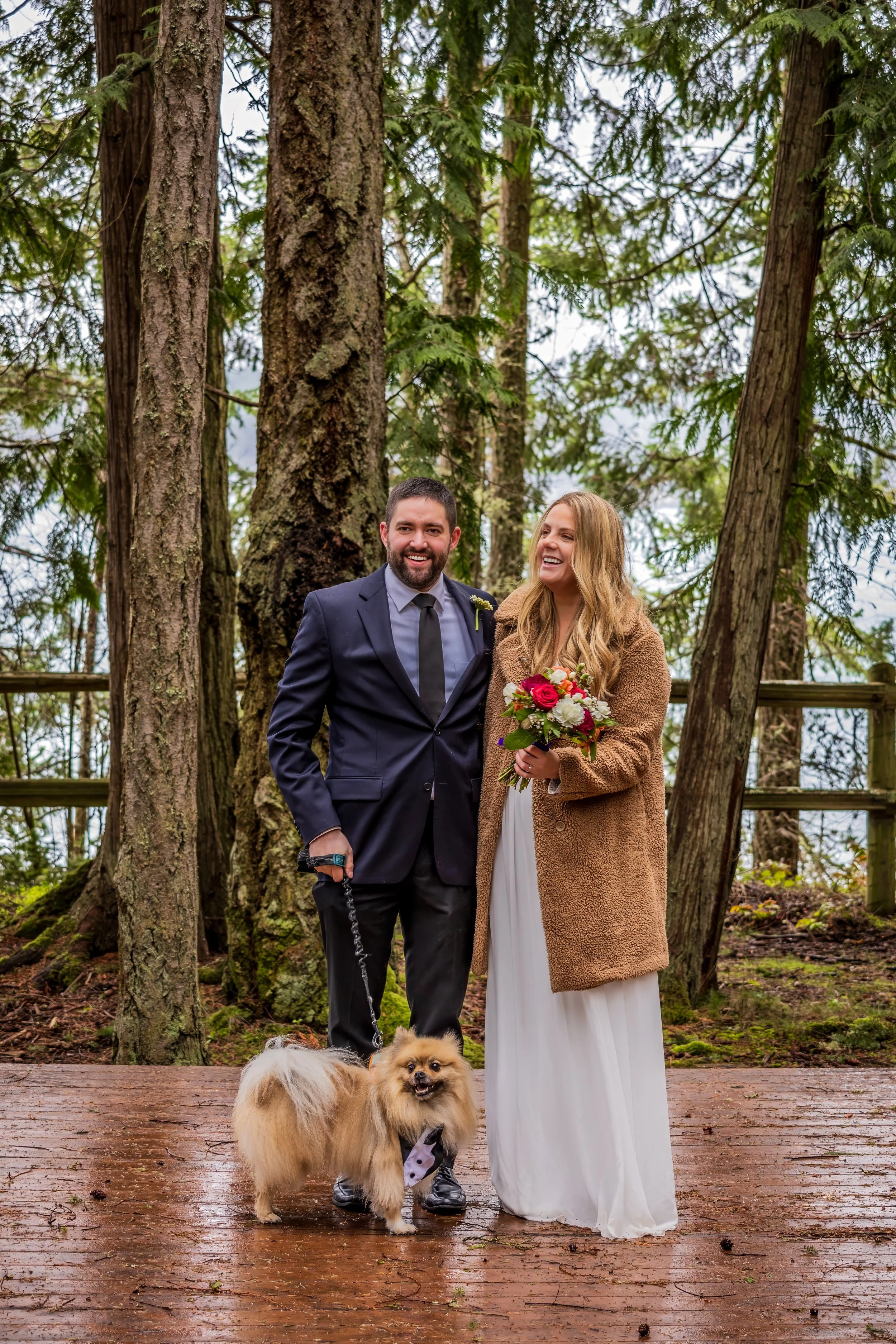 A newlywed couple standing on a wooden platform in a forest, smiling, with a small fluffy dog, dressed with a bow tie, on a leash, in front of them. The woman is holding a bouquet of flowers, and the man is wearing a suit.