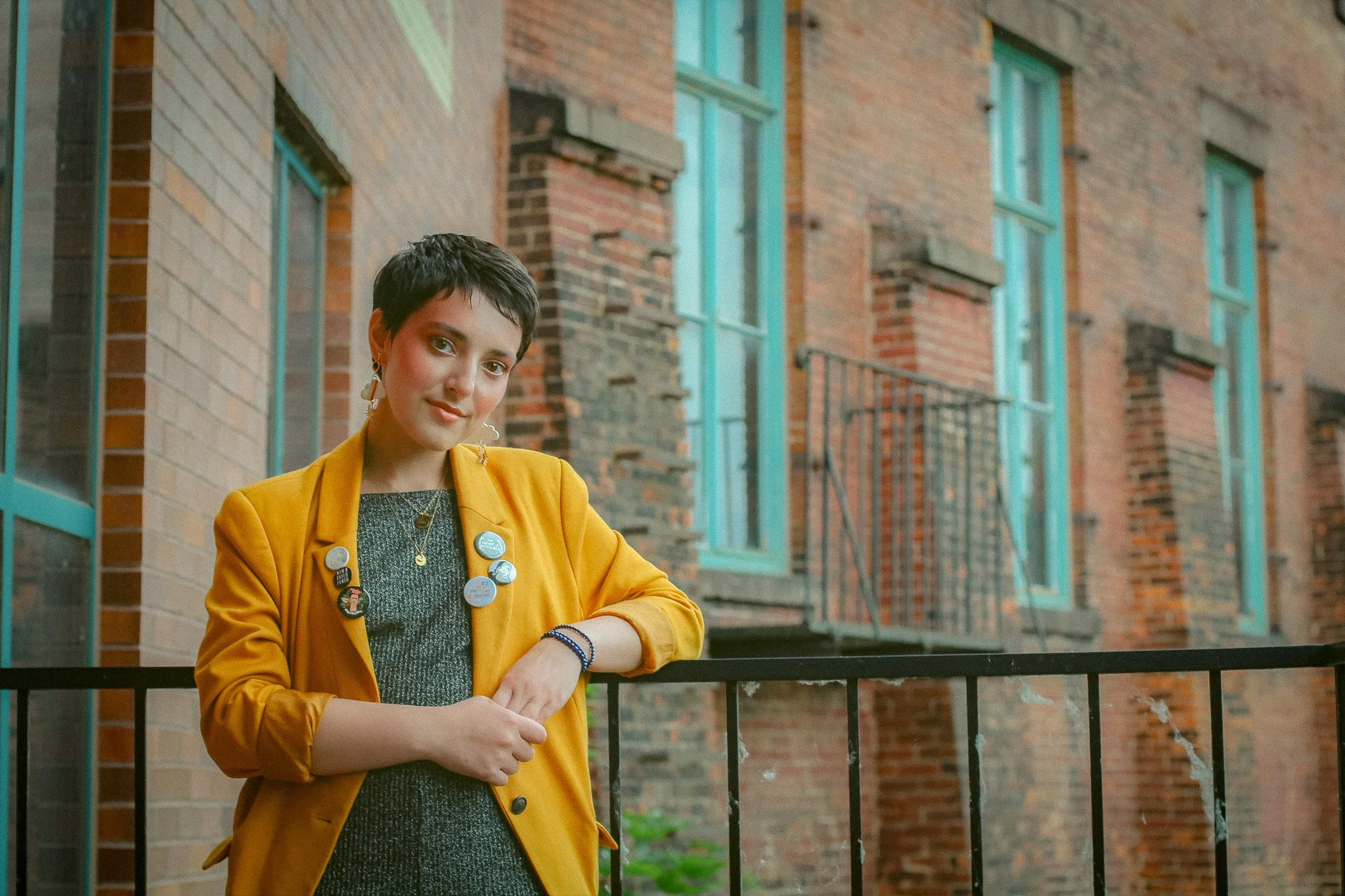 A woman with short dark hair, wearing a mustard yellow blazer with pins and badges, stands outside in front of a brick building with blue window frames, leaning on a black metal fence.