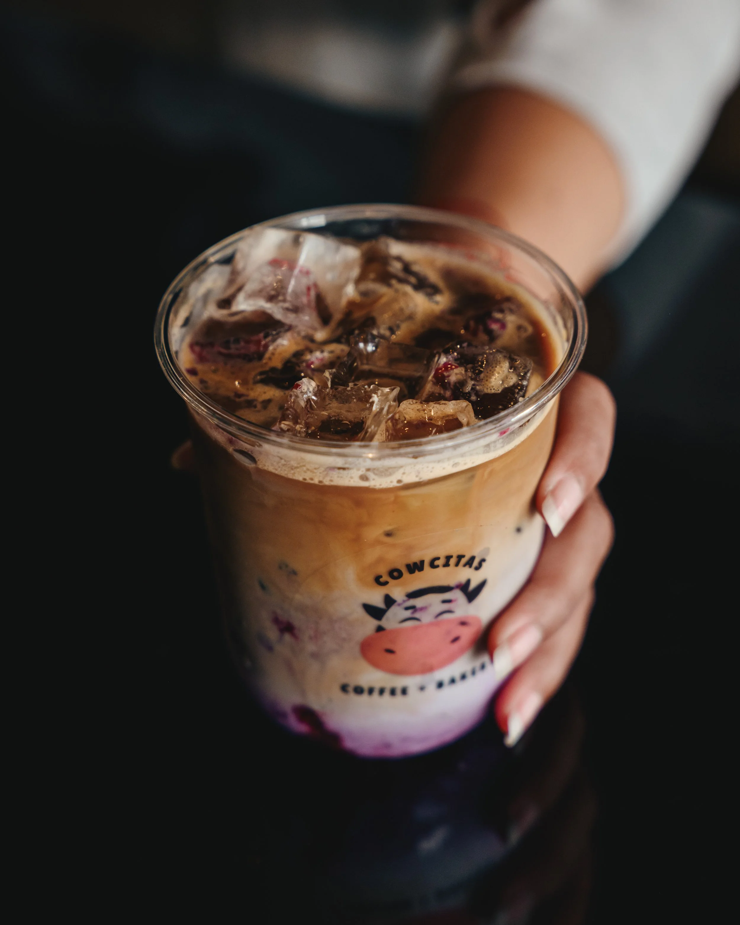 A person holding a plastic cup with iced coffee and ice cubes, featuring a logo with a cartoon cow's face and the text 'COWCIAS.'