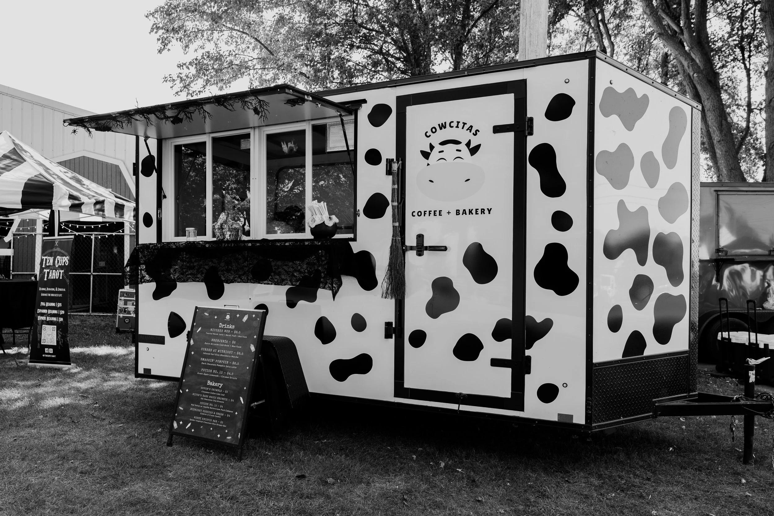 Black and white photo of a cow-themed food truck with the words 'Cowcitas Coffee + Bakery' on the door, parked on grass with other tents and trees in the background.