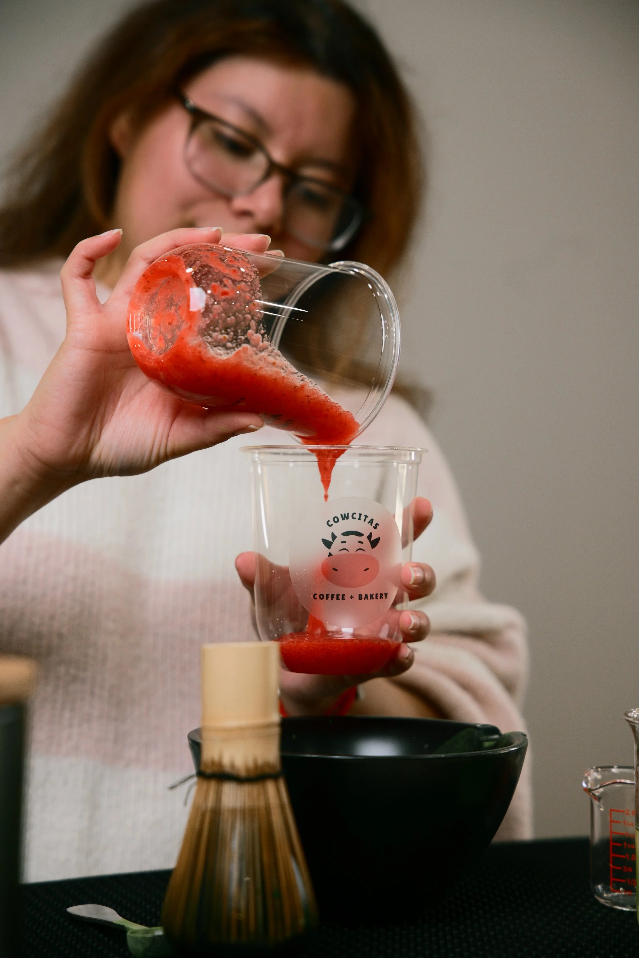 A woman with glasses pouring strawberry puree into a clear cup with a cow mascot logo that says 'COWITAS Coffee + Bakery.' She is in a kitchen or dining area, with a black bowl, whisk, and measuring cup nearby.