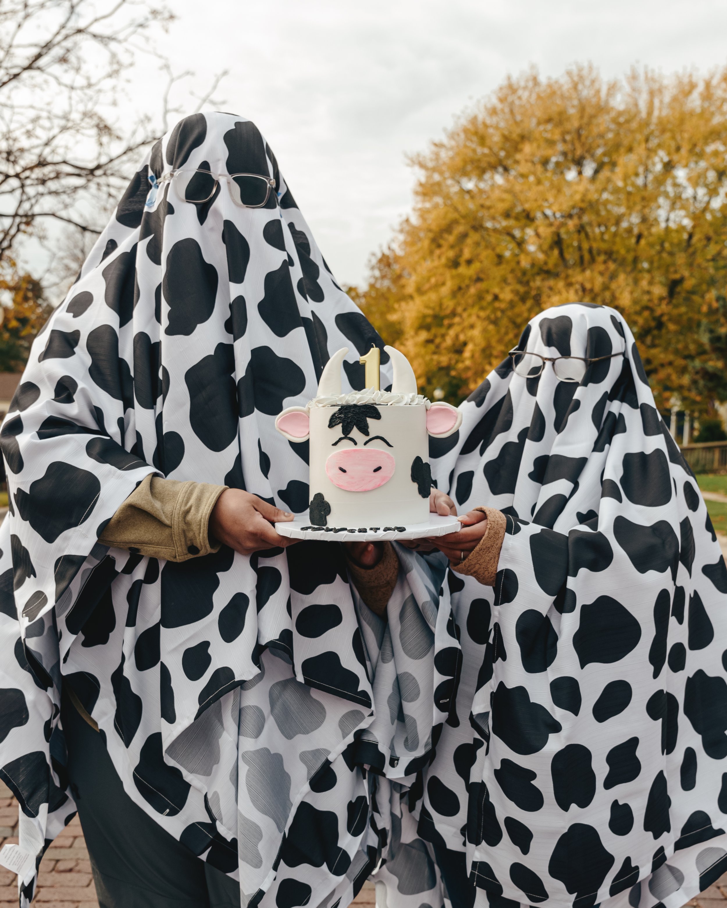 Two people dressed in cow-themed costumes holding a cake with a cow face design, outdoors with autumn trees in the background.