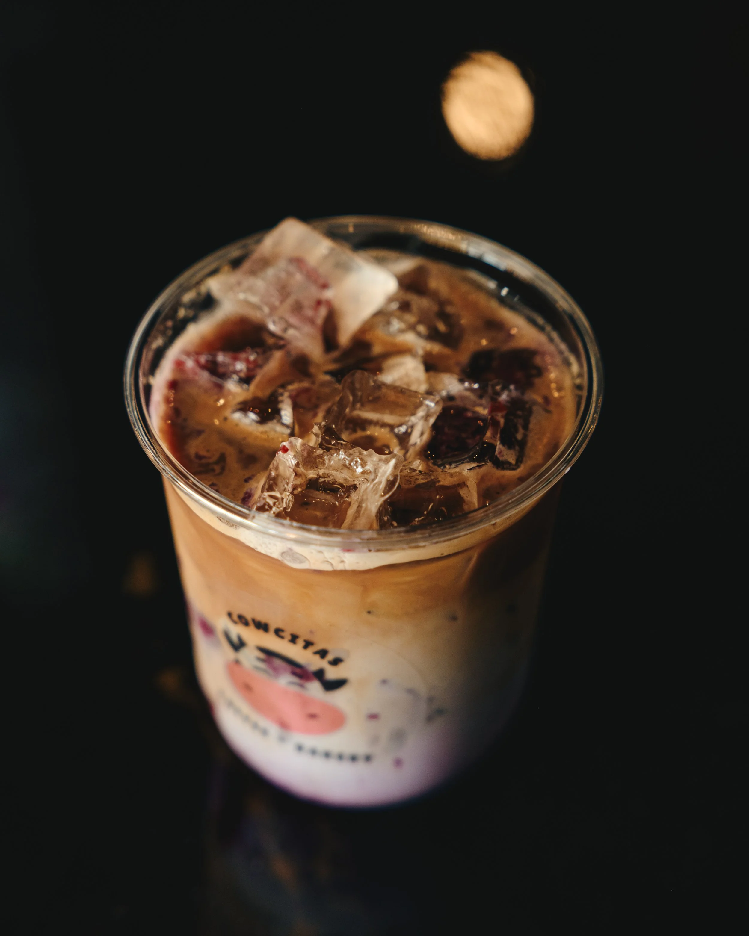 Ice coffee beverage with ice cubes in a clear cup with a logo, on a dark surface with a blurred light reflection.