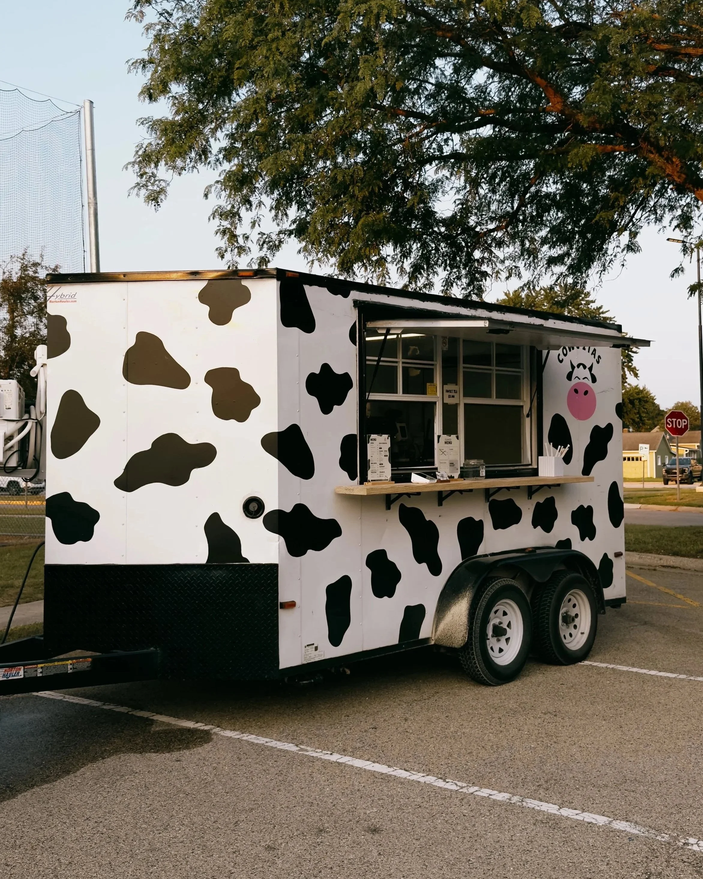 A cow-themed food trailer parked in a parking lot with trees and a stop sign in the background, featuring black and white cow spots and a pink cow face illustration on the side.