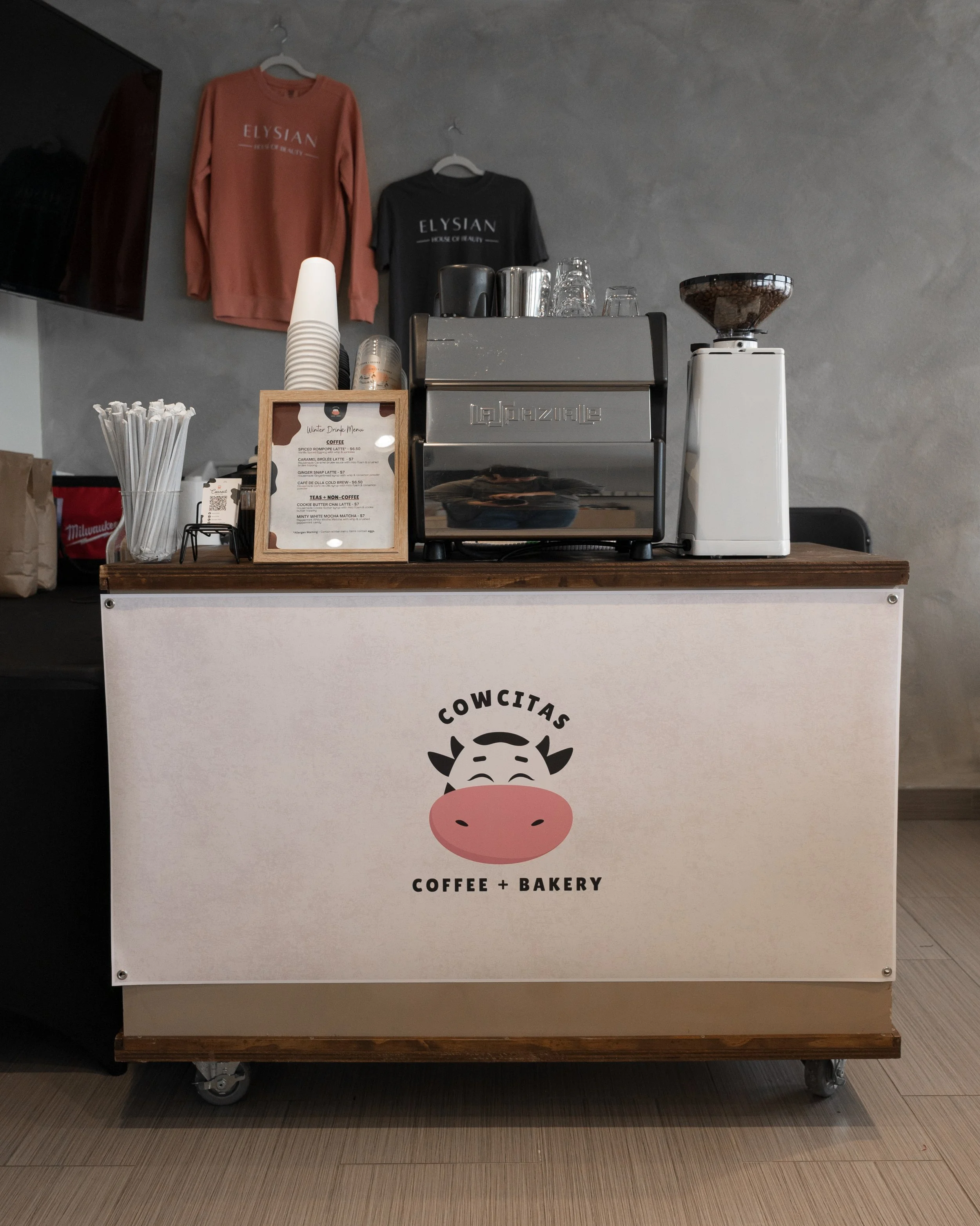 A coffee and bakery cart with a cow logo and the words "Cowcitas Coffee + Bakery," featuring a coffee machine, grinder, cups, straws, and T-shirts hanging on the wall behind.