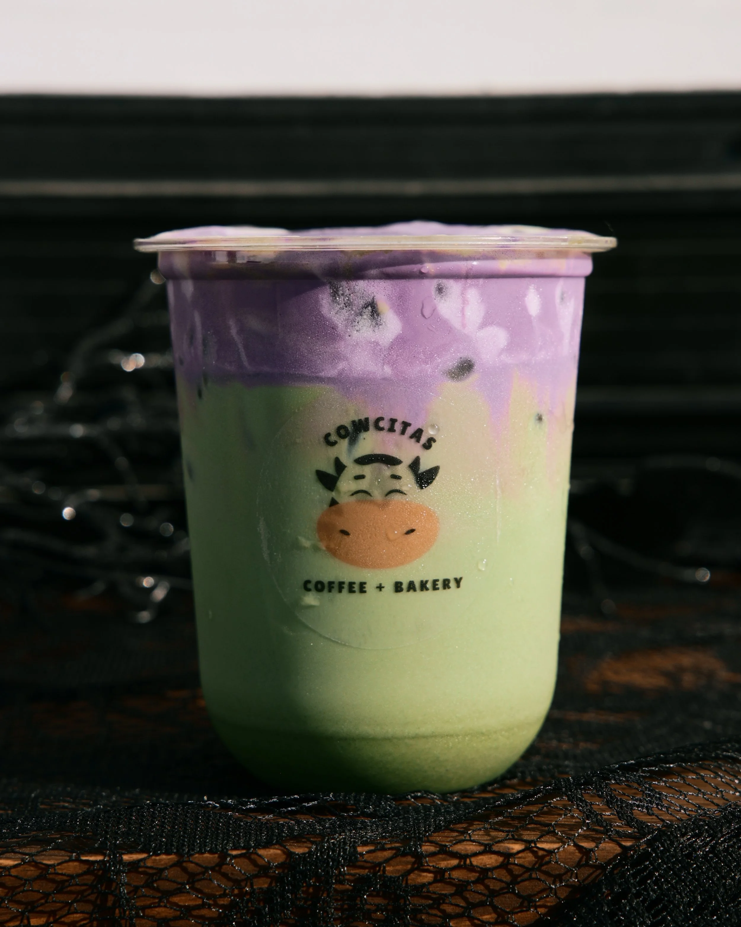 Close-up of a colorful coffee cup with a cow face design and the text "CowCityas Coffee + Bakery" on it, resting on a dark textured surface.