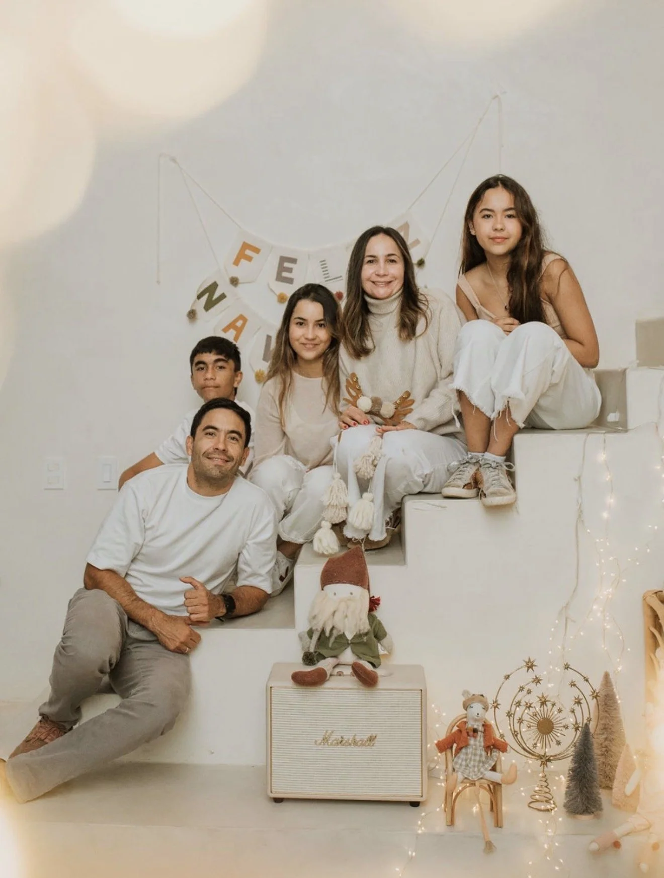Family of six posing on white stairs decorated with Christmas ornaments, lights, and holiday decorations, with a "FELIZ NAVIDAD" banner in the background.