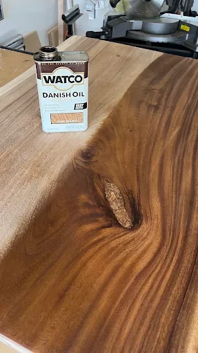 A bottle of Watco Danish Oil on a wooden table with a natural wood finish and a knot in the wood.