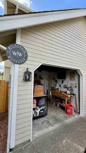 View of a garage with a partial view of the house exterior showing beige siding, an awning, and a circular sign that says 'Wildgrain Foodshop'. Inside the garage are shelves with various items, a workbench, and storage containers.