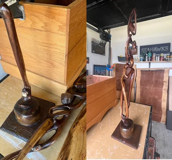 A tall, abstract wooden sculpture of intertwined figures on a rectangular base, located in a garage or workshop.