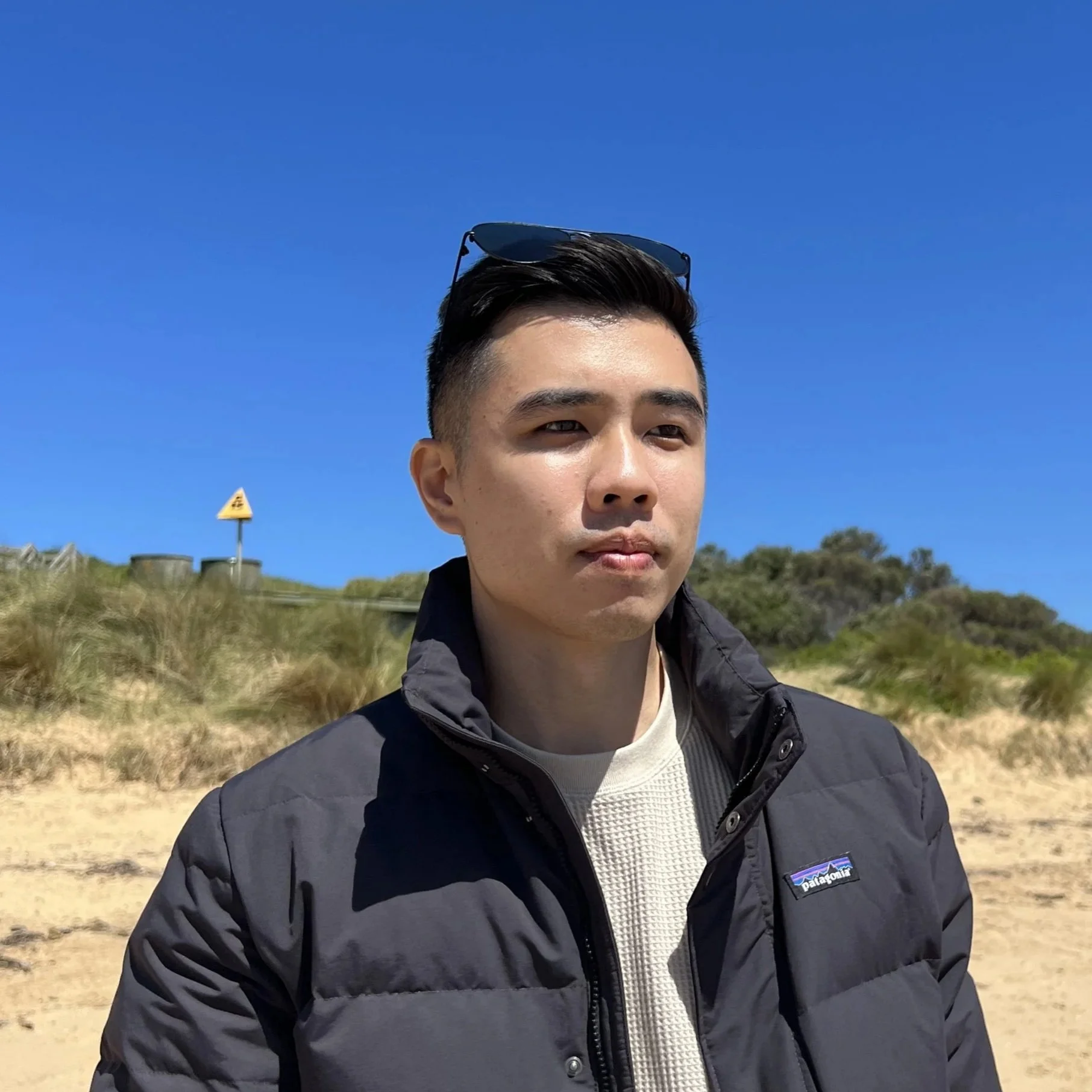 Young man standing on a sandy beach with dunes and shrubs, wearing a black Patagonia jacket and sunglasses on his head, under a clear blue sky.